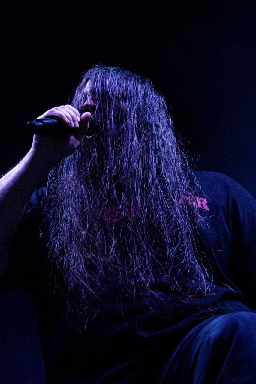 A person with long, wet hair holding a microphone near their face, under dark, purple-lit lighting.
