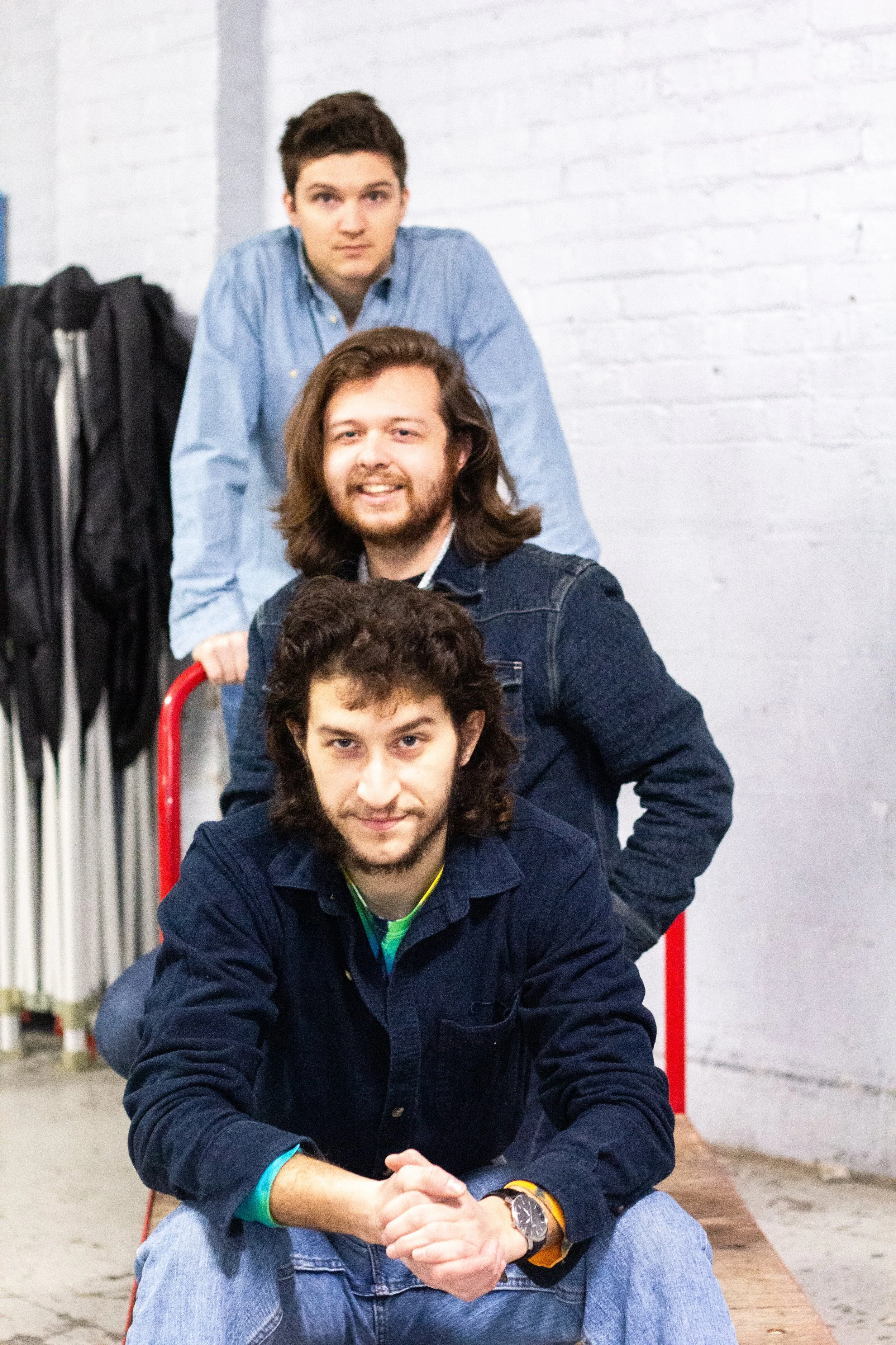 Four men of varying ages and hairstyles stacking on top of each other, seated on chairs, with a white brick wall and some equipment in the background.