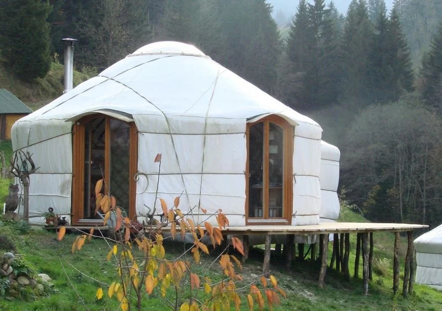 A yurt made of white fabric with wooden doors, elevated on wooden stilts in a rural, forested area.