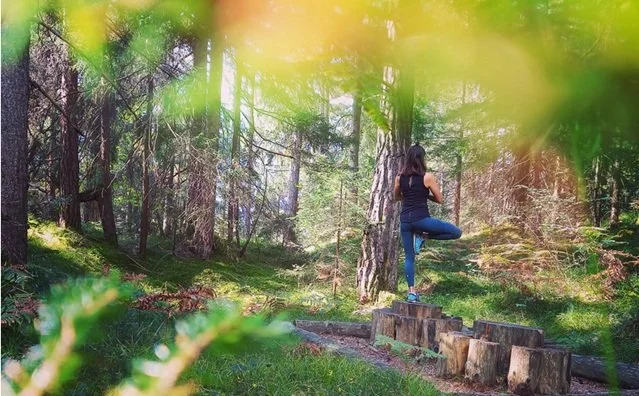 A woman practicing yoga outdoors in a forest, standing on one leg with hands in a prayer position.