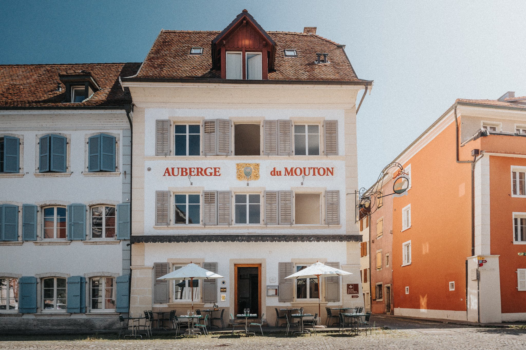 Bâtiment coloré avec un restaurant appelé 'Auberge du Mouton', terrasse extérieure avec chaises, tables et parasols, au bord d'une rivière, avec des fenêtres à volets.