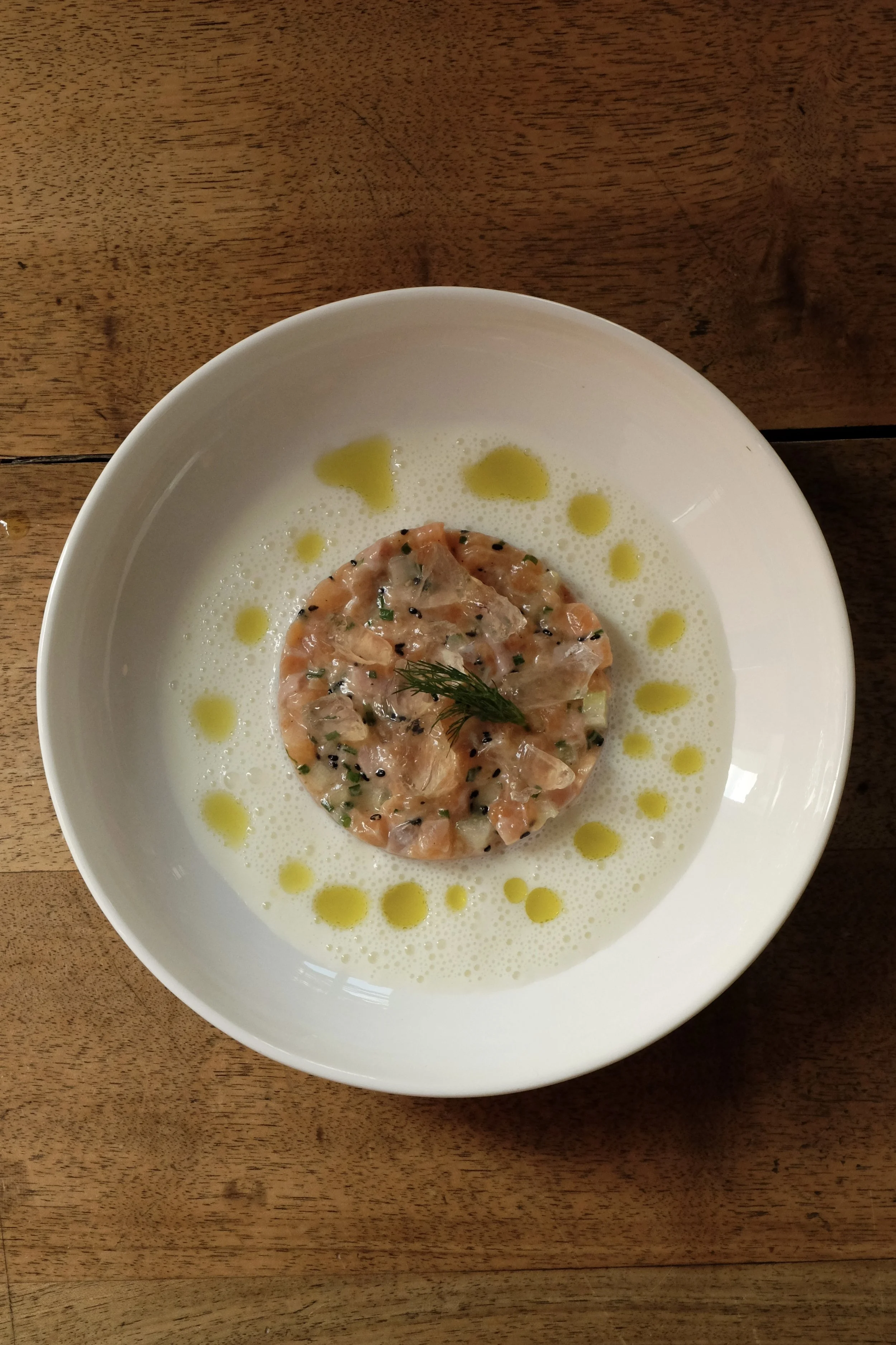 Carpaccio de saumon avec une garniture d'aneth, entouré d'huile d'olive sur une assiette blanche, sur une table en bois.