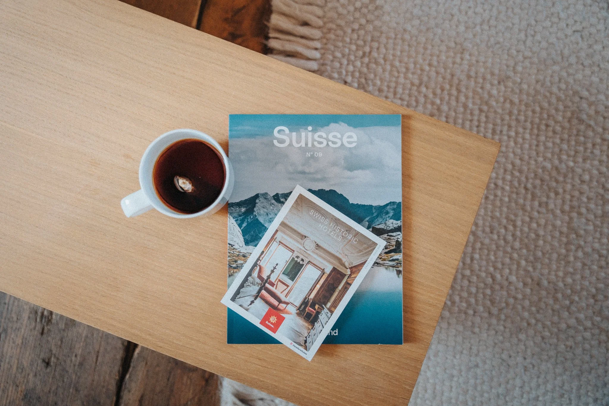 Une tasse de café noir à côté d'un magazine intitulé "Suisse" avec une photo de montagnes et un petit flyer représentant un intérieur avec des meubles, posé sur une table en bois.