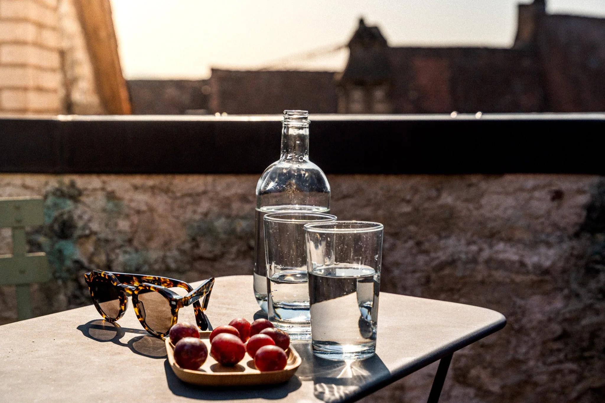 Une table avec des lunettes de soleil, un panier de fruits, deux verres d'eau et une bouteille en verre, en extérieur avec une maison en pierre en arrière-plan.