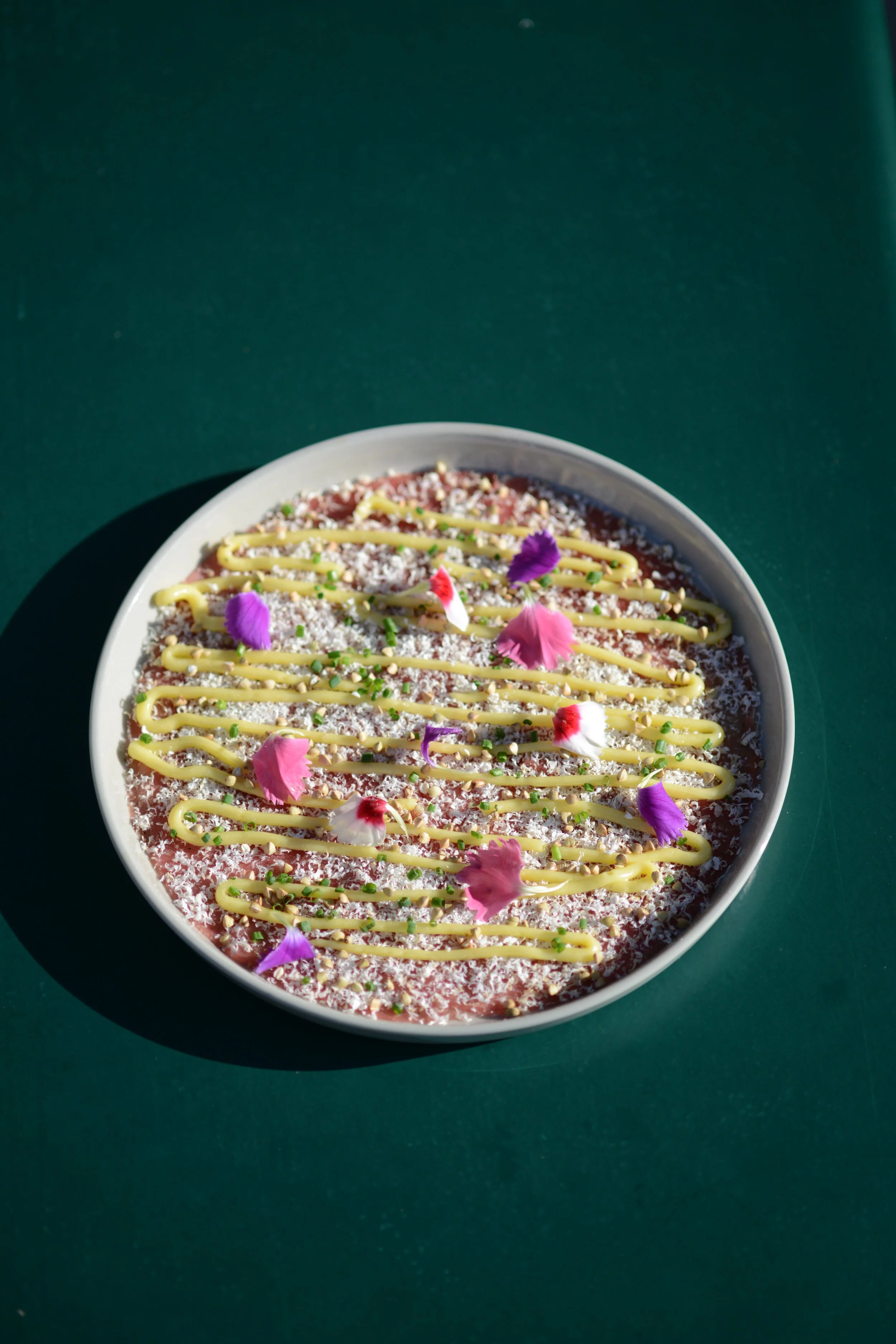Une pâtisserie ronde décorée avec des fleurs, des graines vertes, de la poudre blanche, du chocolat en filets et de la sauce jaune.