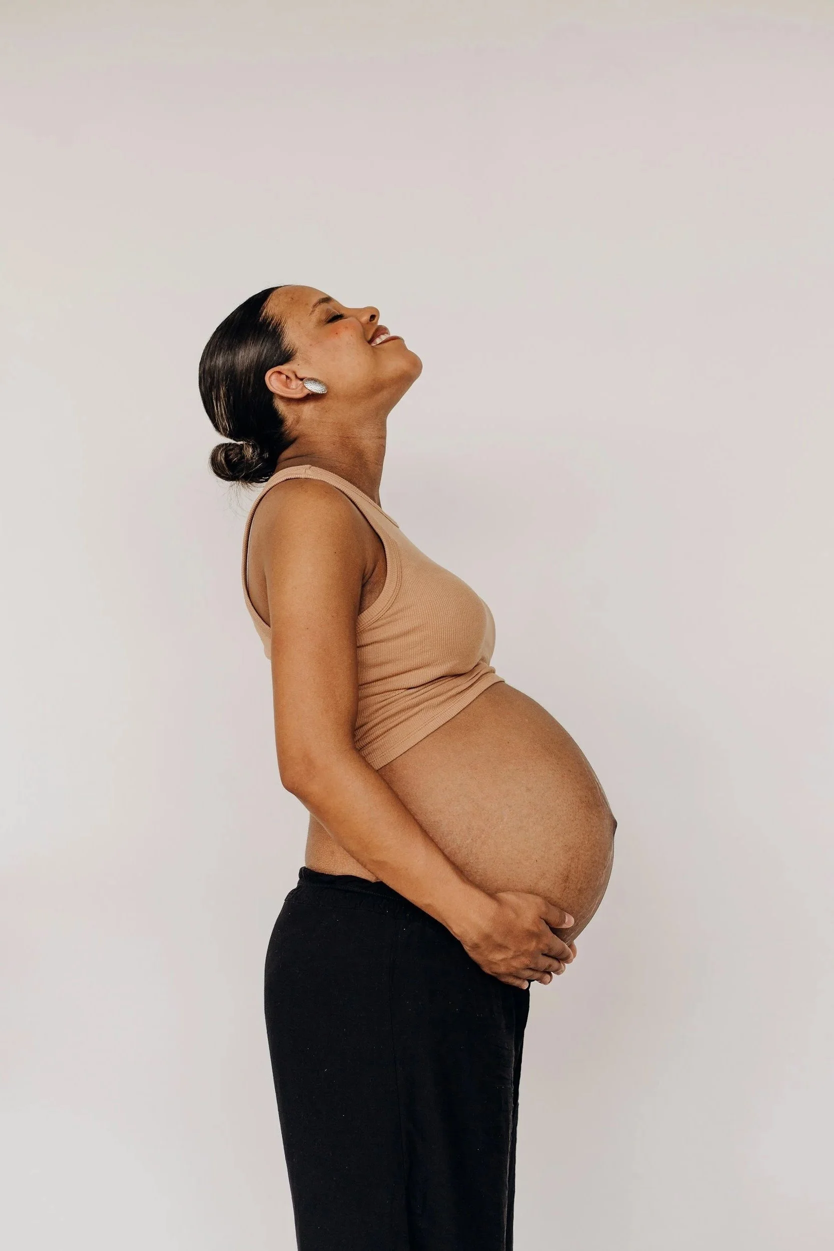 Pregnant woman with dark hair in a bun, wearing a beige tank top and black pants, standing sideways with her head tilted back and smiling, against a plain white background.