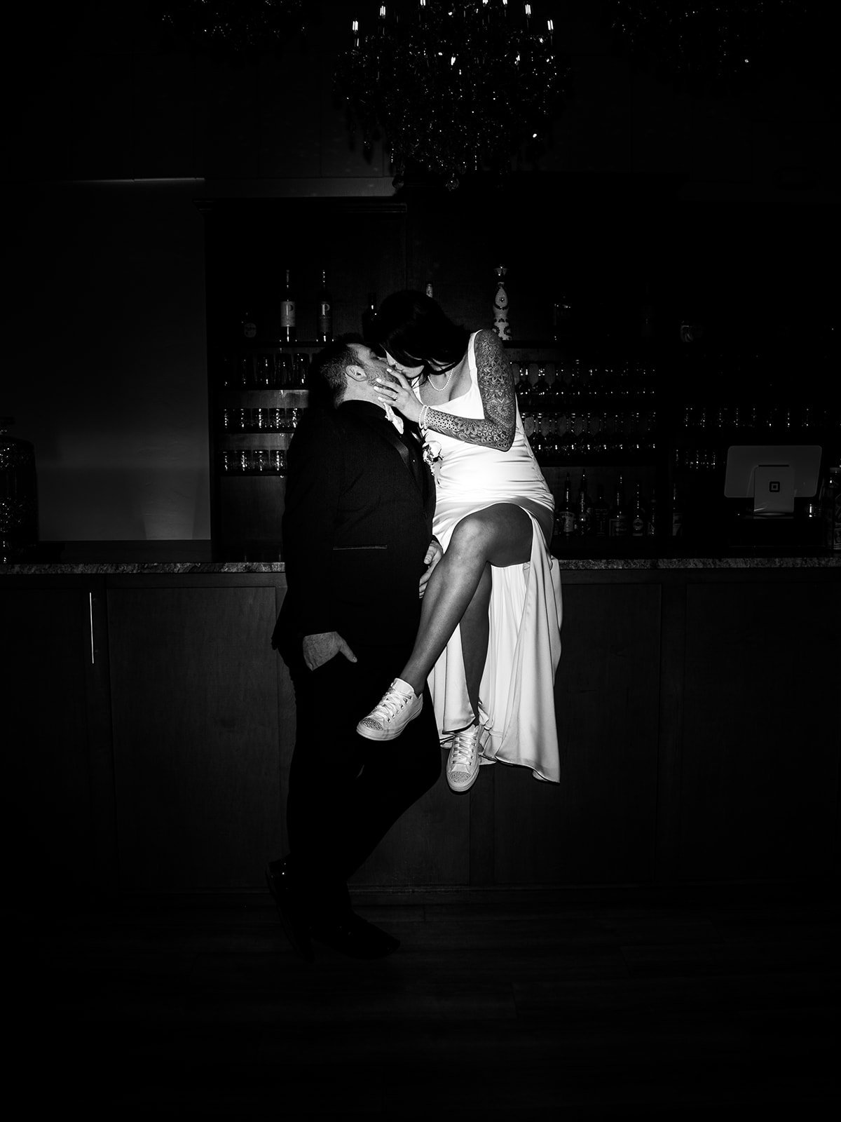 A black-and-white photo of a man in a tuxedo holding a woman in a white dress and sneakers, with her leg lifted, sitting on the bar of the Ledgecrest Reserve wedding venue in Green Bay, Wisconsin.