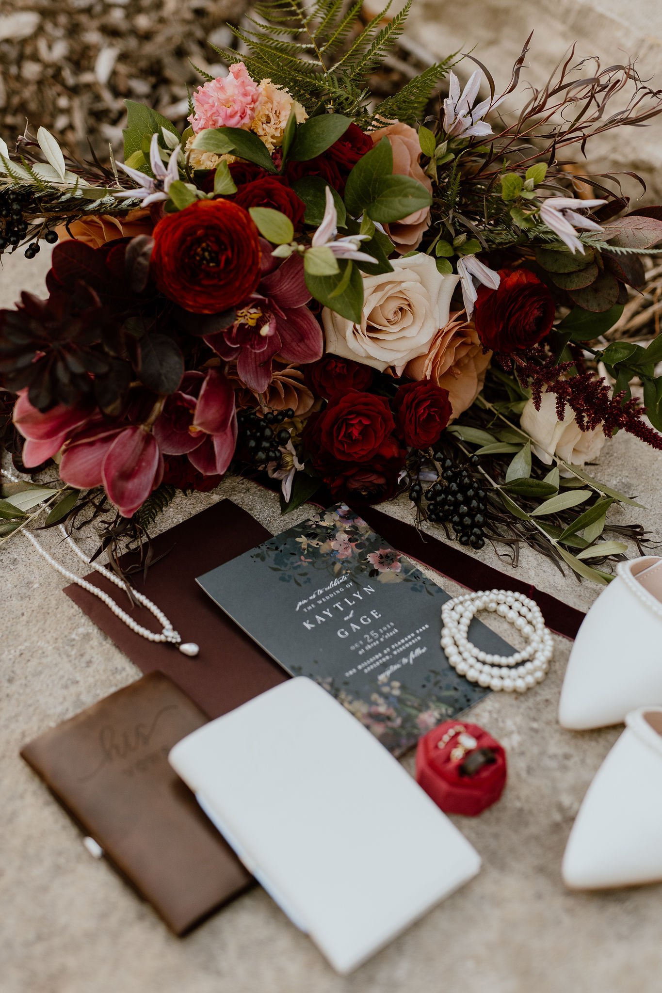 Wedding invitation surrounded by a pearl necklace, a small red ring box, and a floral bouquet with roses and greenery.