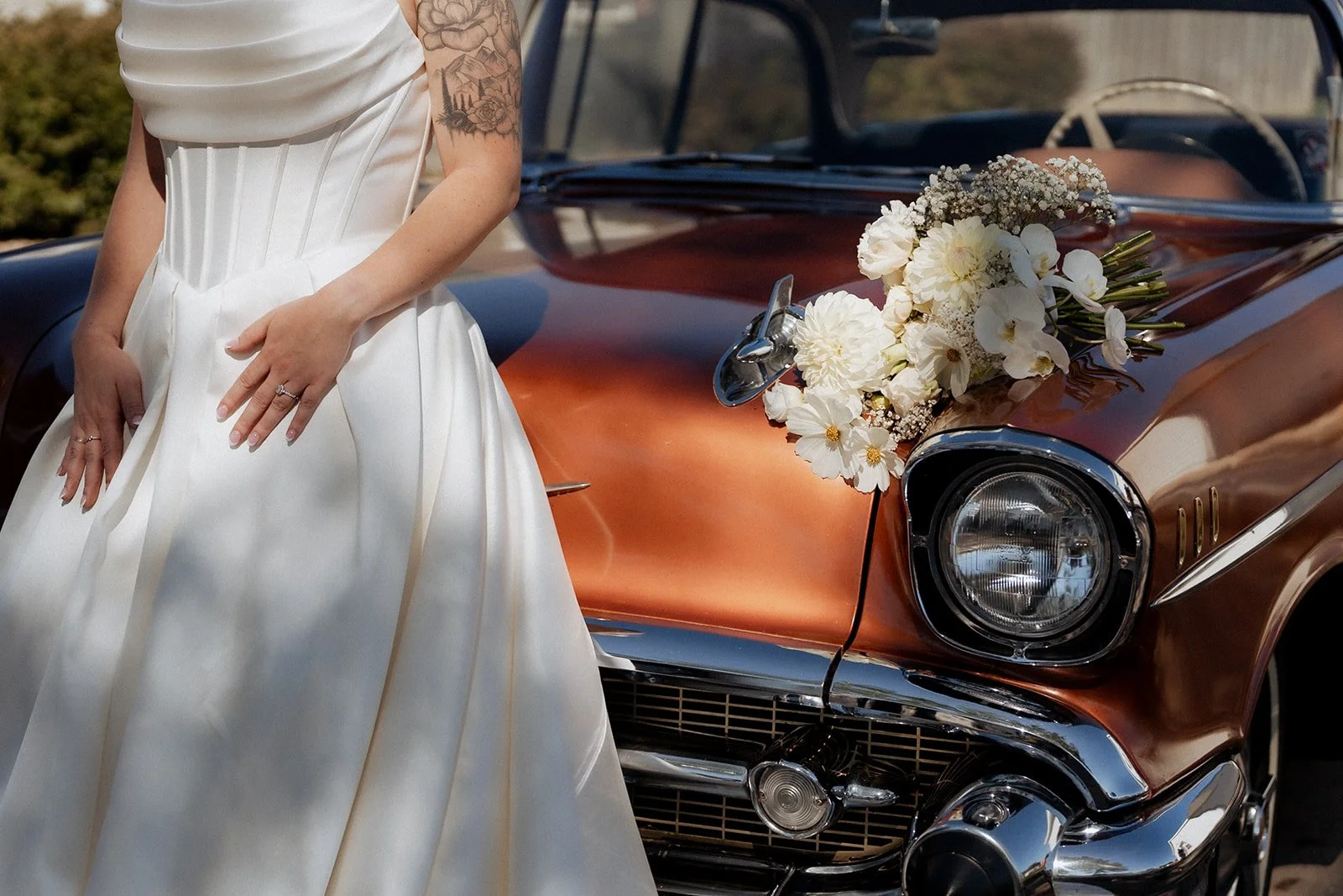 Part of a bride wearing a white dress and rings, standing next to a vintage orange car with a bouquet of white flowers on its hood in Green Bay, Wisconsin.