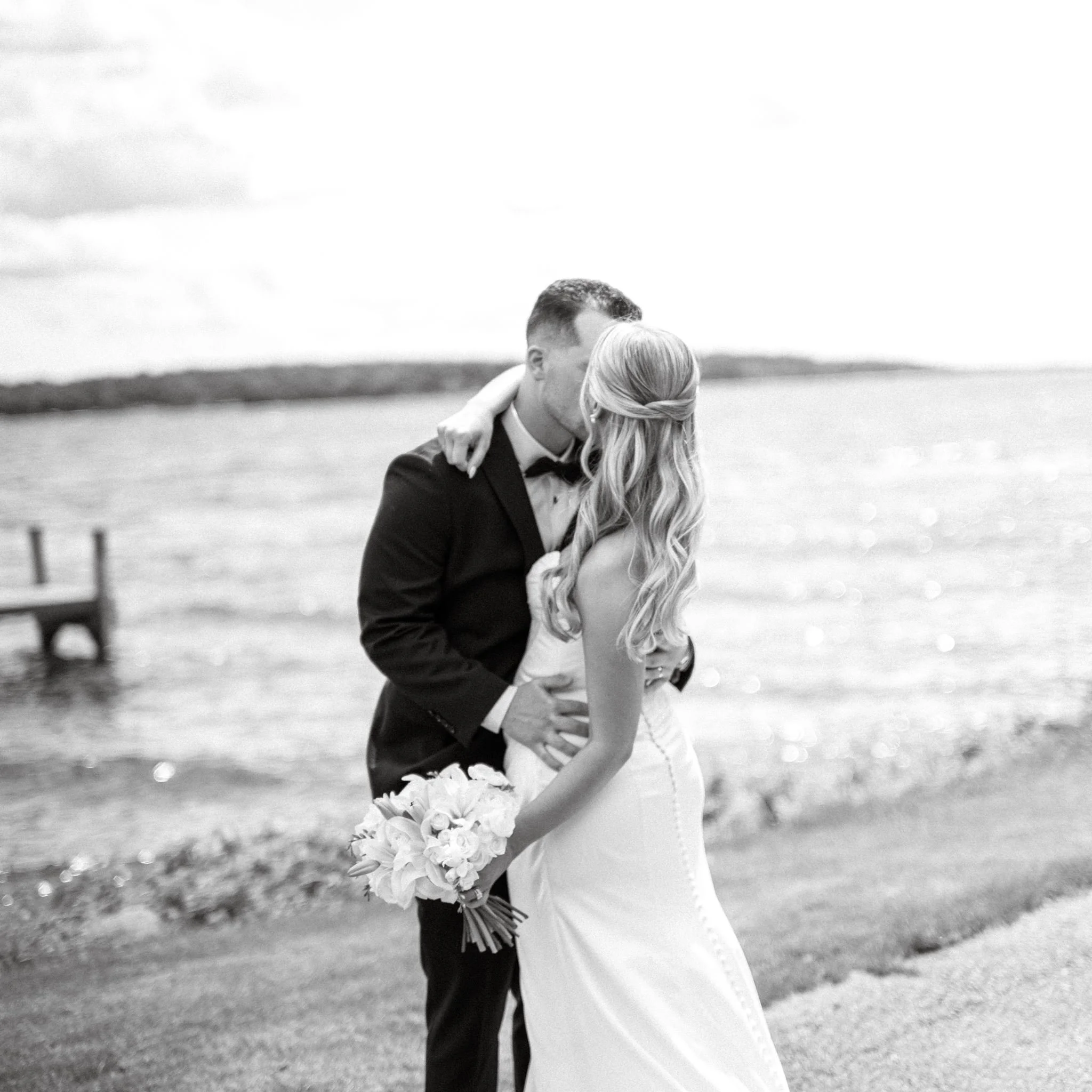 A couple, dressed in wedding attire, sharing a kiss outdoors with a bouquet of flowers in the foreground. The background is Green Lake, Wisconsin.