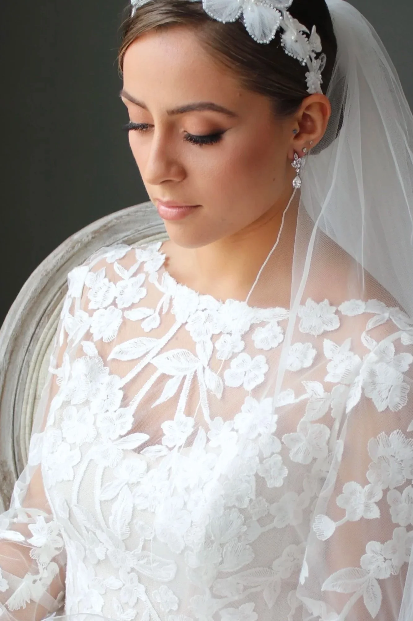 A bride in a white lace wedding dress with floral embroidery, wearing a white floral headband, veil, and elegant earrings, sitting with eyes closed.