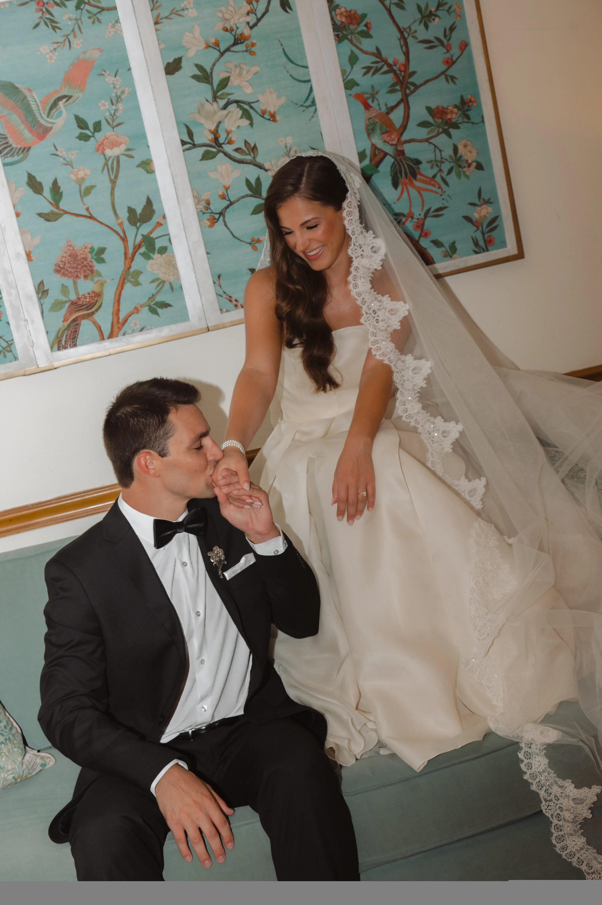 A bride in a wedding dress and veil sitting on a sofa, holding hands with a groom in a tuxedo who is kneeling and kissing her hand, in front of decorative Asian-style painting with birds and flowers. Morgan Parker Photography