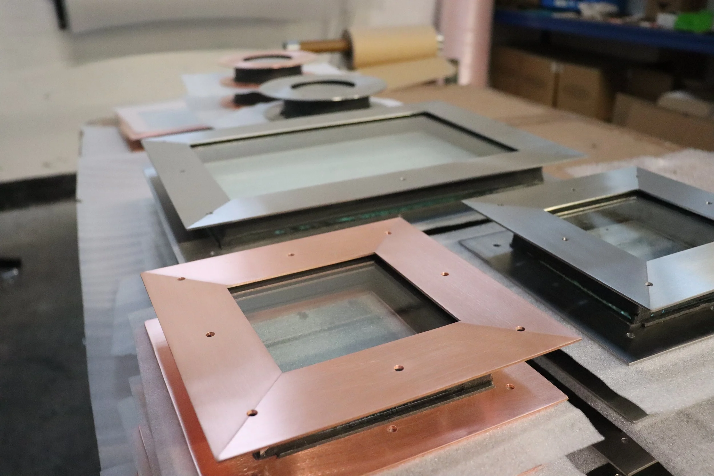 Industrial metal frames with glass inserts, some in copper and others in black, placed on a workbench. Additional materials and tools are visible in the background, indicating a workshop setting.