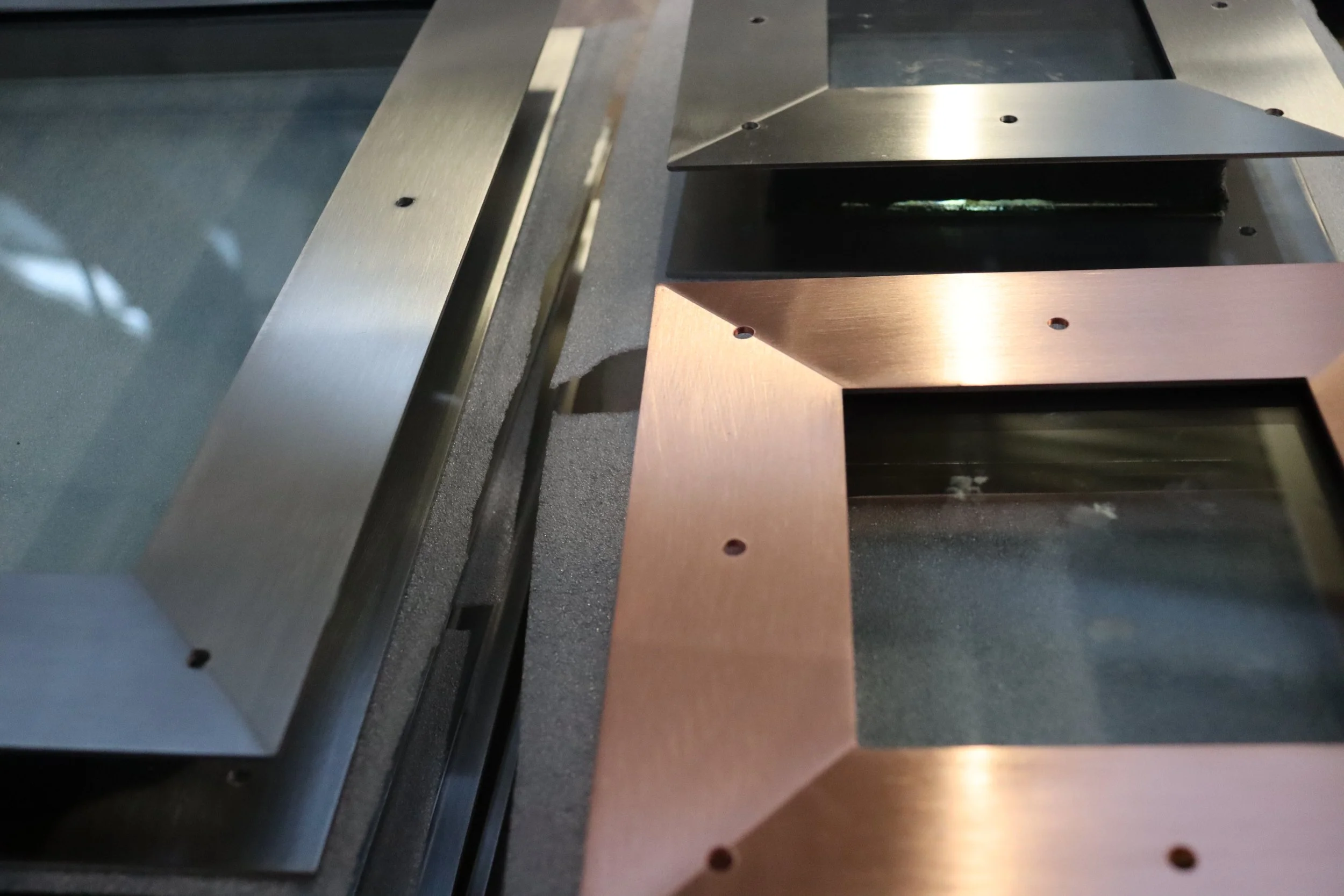 Close-up of metallic rectangular frames, one silver and one copper-colored, with drilled holes, set on a dark textured surface.