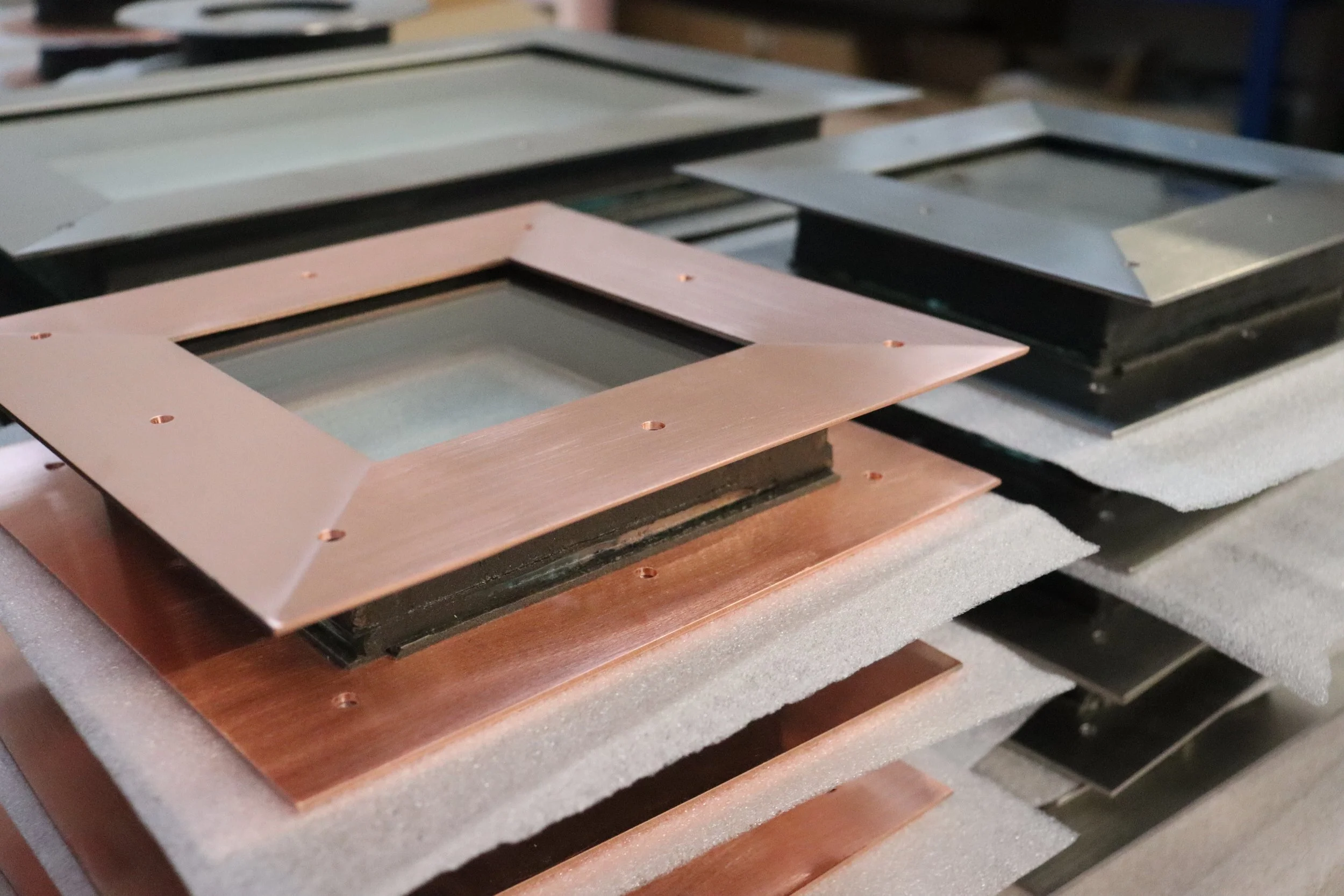Close-up of stacked metal and copper frames with glass inserts, placed on foam padding in a workshop.