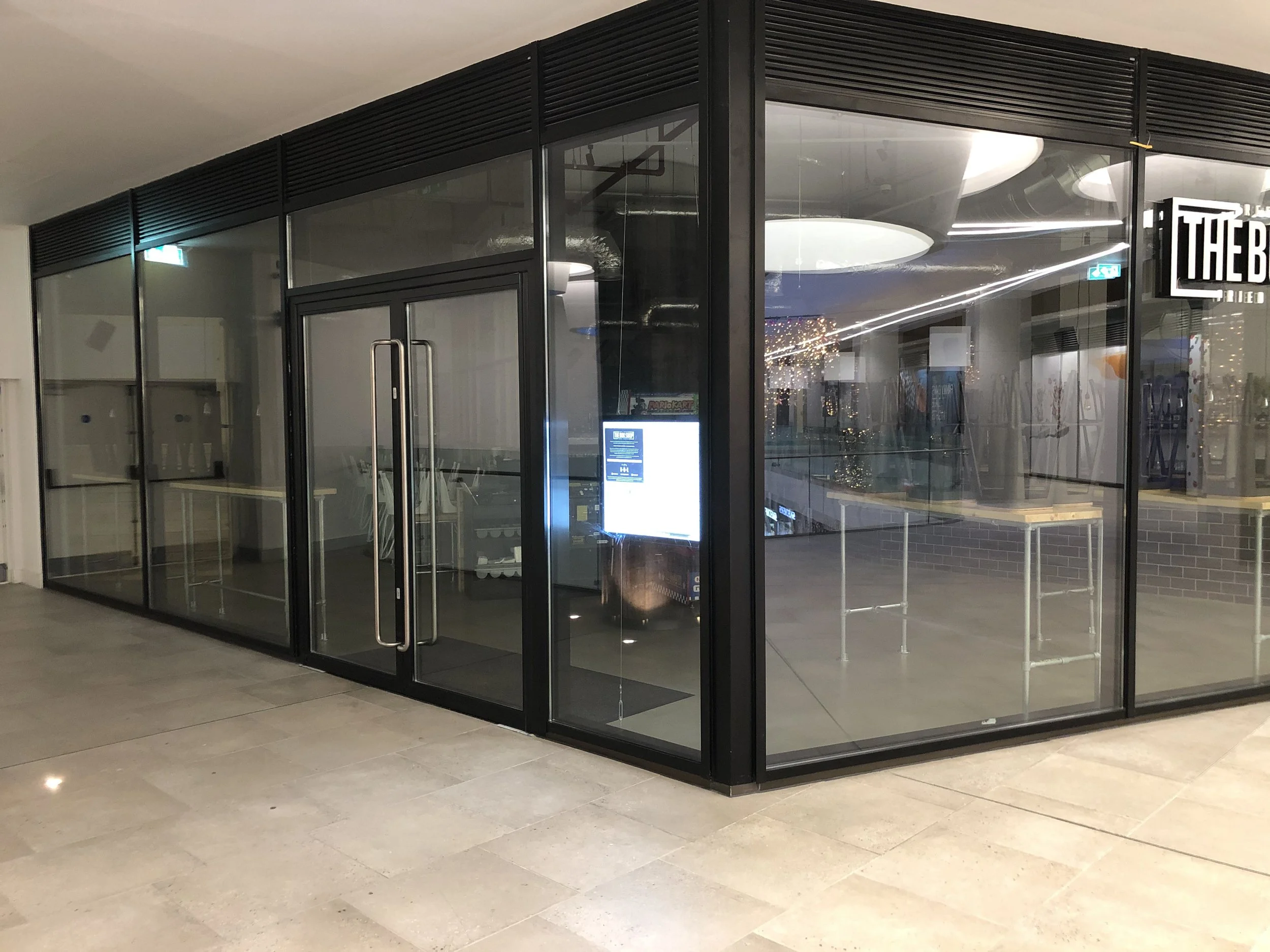 Empty glass storefront with black framing and rolled tables inside, illuminated by ceiling lights, sign partially visible on the right, located at an indoor shopping mall or commercial area.