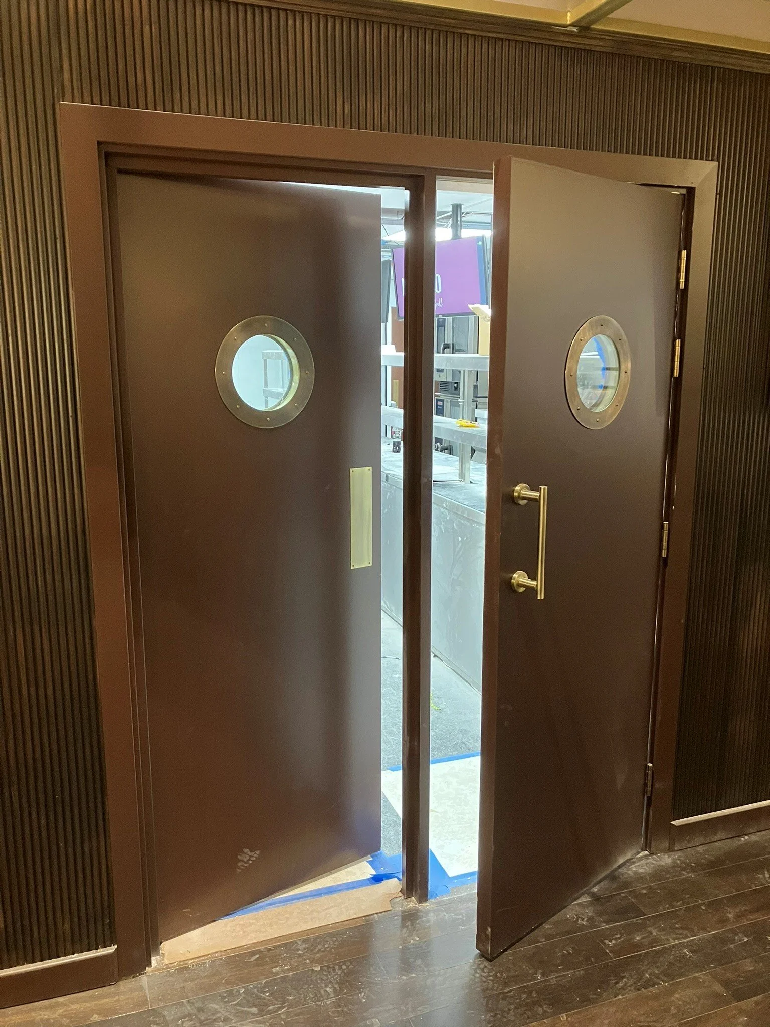 Open double brown doors with circular windows, leading to a room with a purple sign, metal shelving, and various equipment, in a building with dark wood flooring.