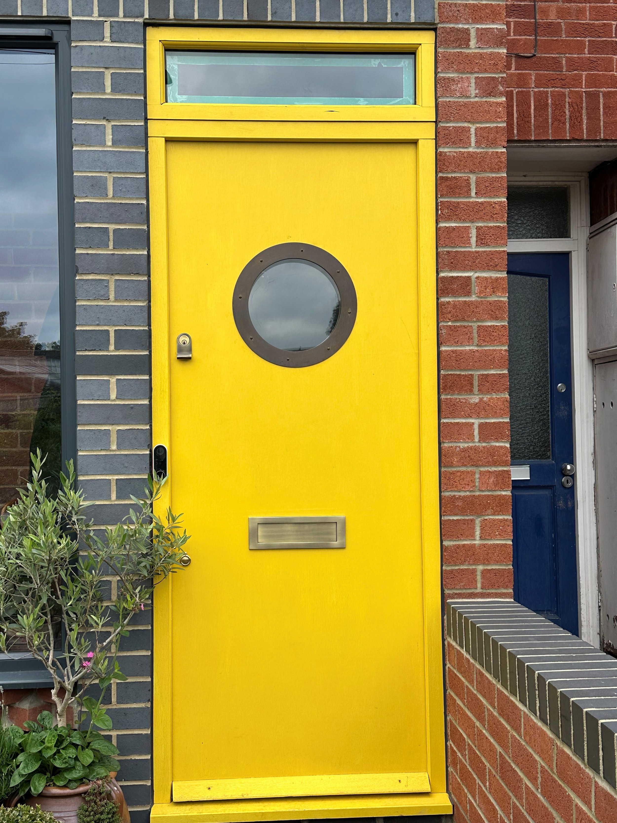 Bright yellow door with a circular window and a horizontal mail slot, set between brick and dark-colored walls.