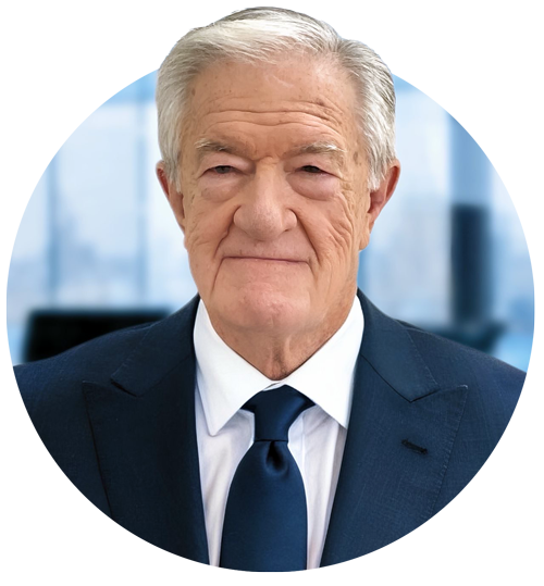 An elderly man with gray hair wearing a dark blue suit, white shirt, and blue tie, standing in a modern office with large windows in the background.
