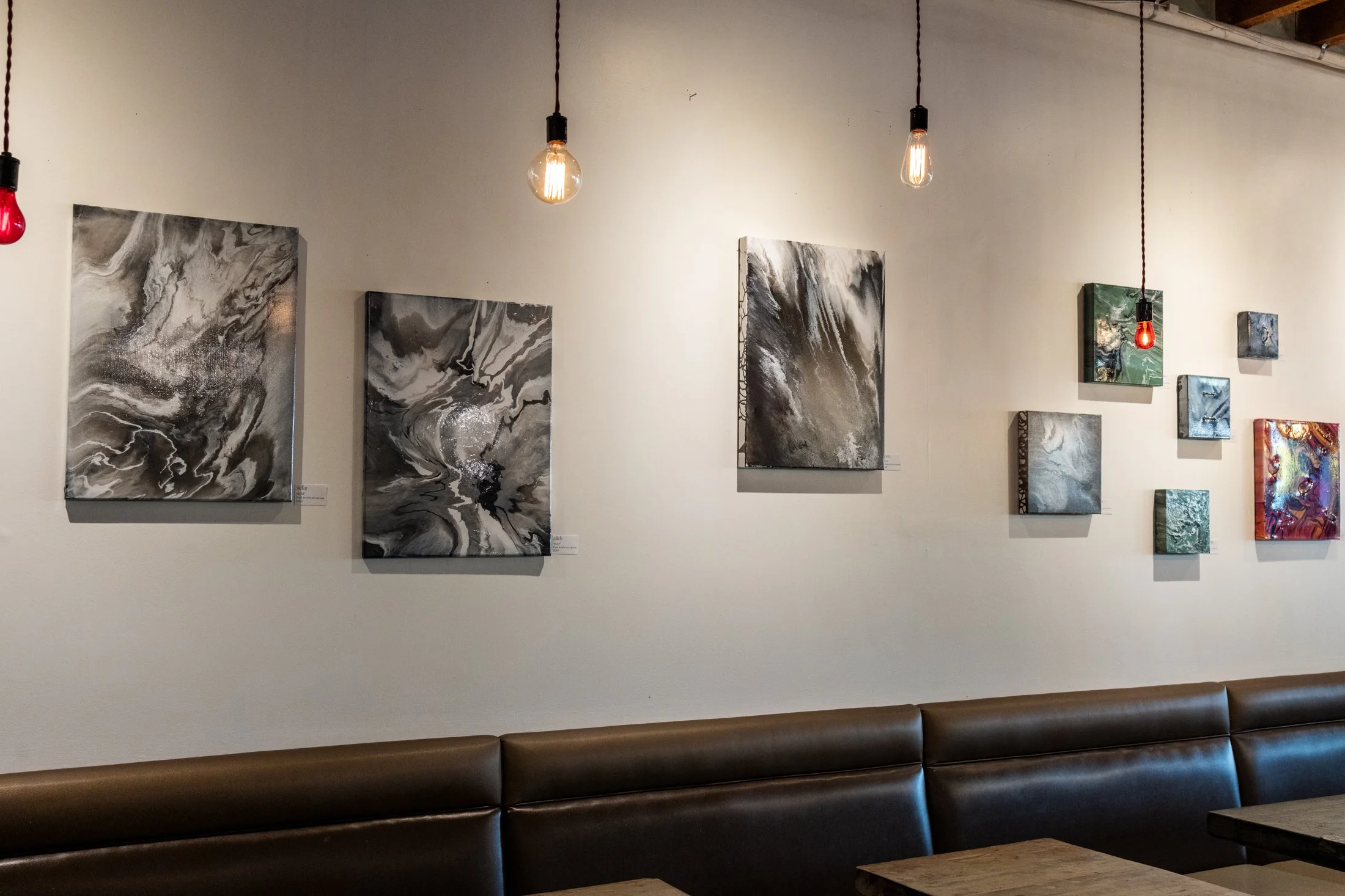 Gallery wall with abstract black, white, and gray paintings illuminated by hanging Edison-style light bulbs in a modern interior.
