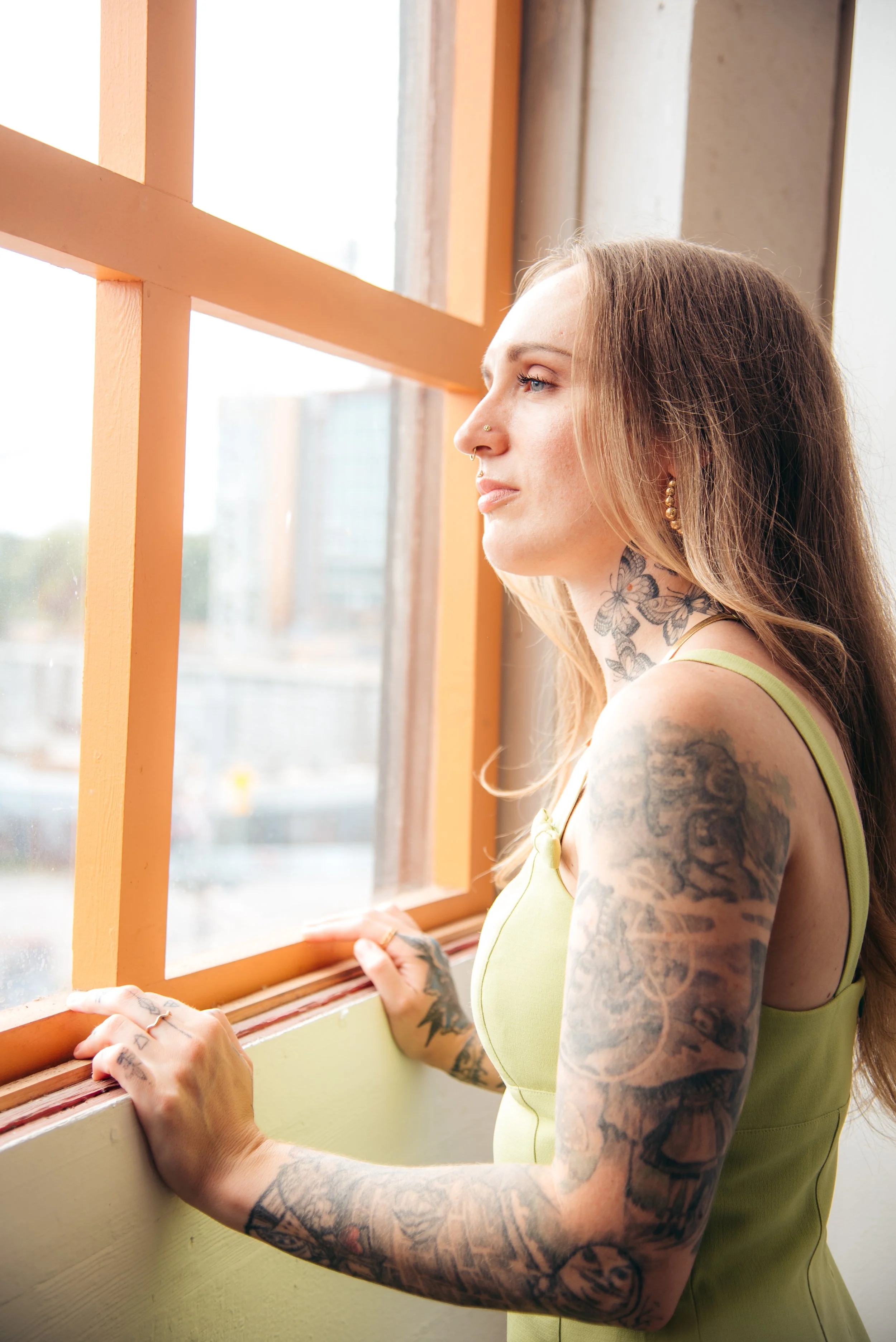 A woman with long hair, tattoos on her arms and neck, wearing earrings, looking out of a window with orange frames, during daytime.