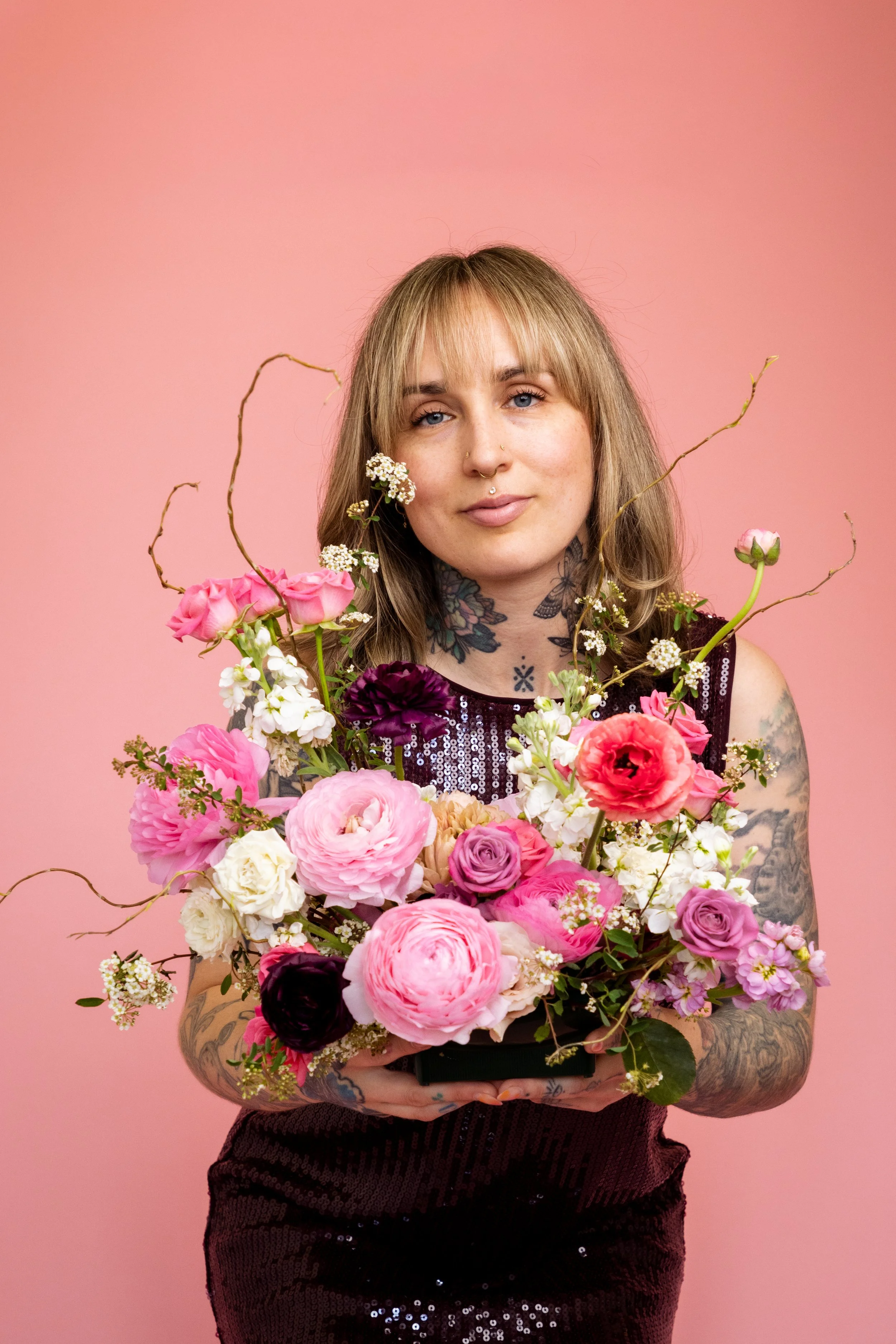 A young woman with blonde hair, tattoos, and piercings holding a large bouquet of pink, white, purple, and burgundy flowers against a pink background.