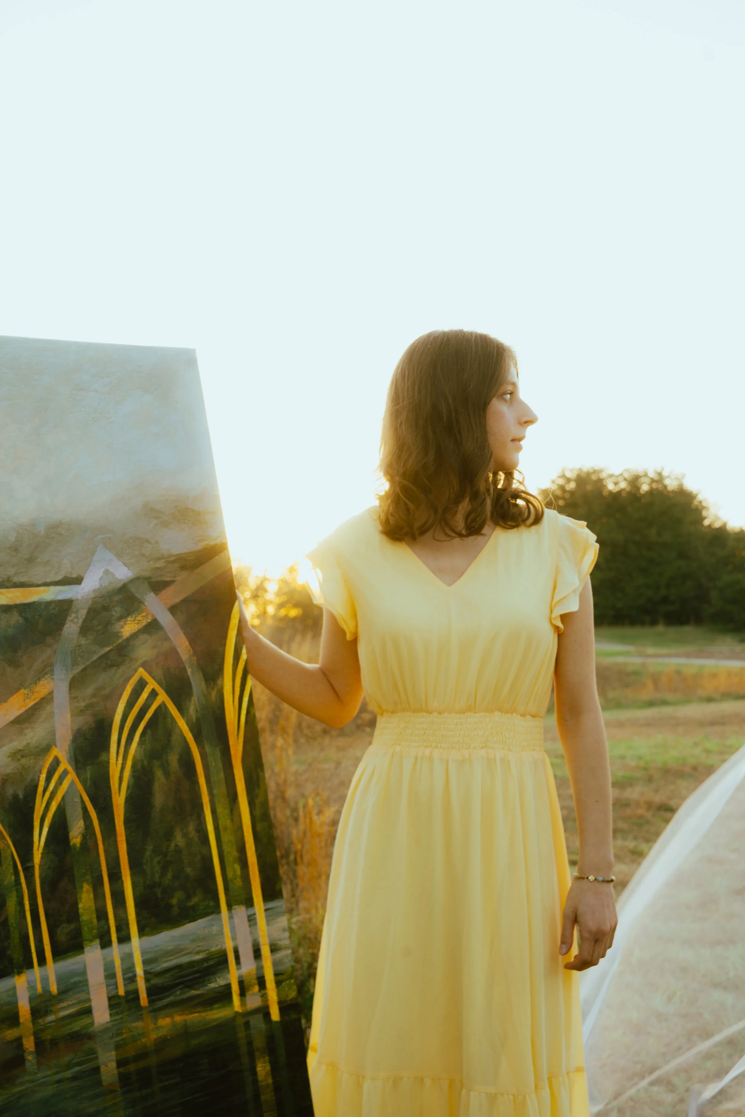 A woman in a yellow dress standing outdoors at sunset, looking to her right, with artworks leaning against her.