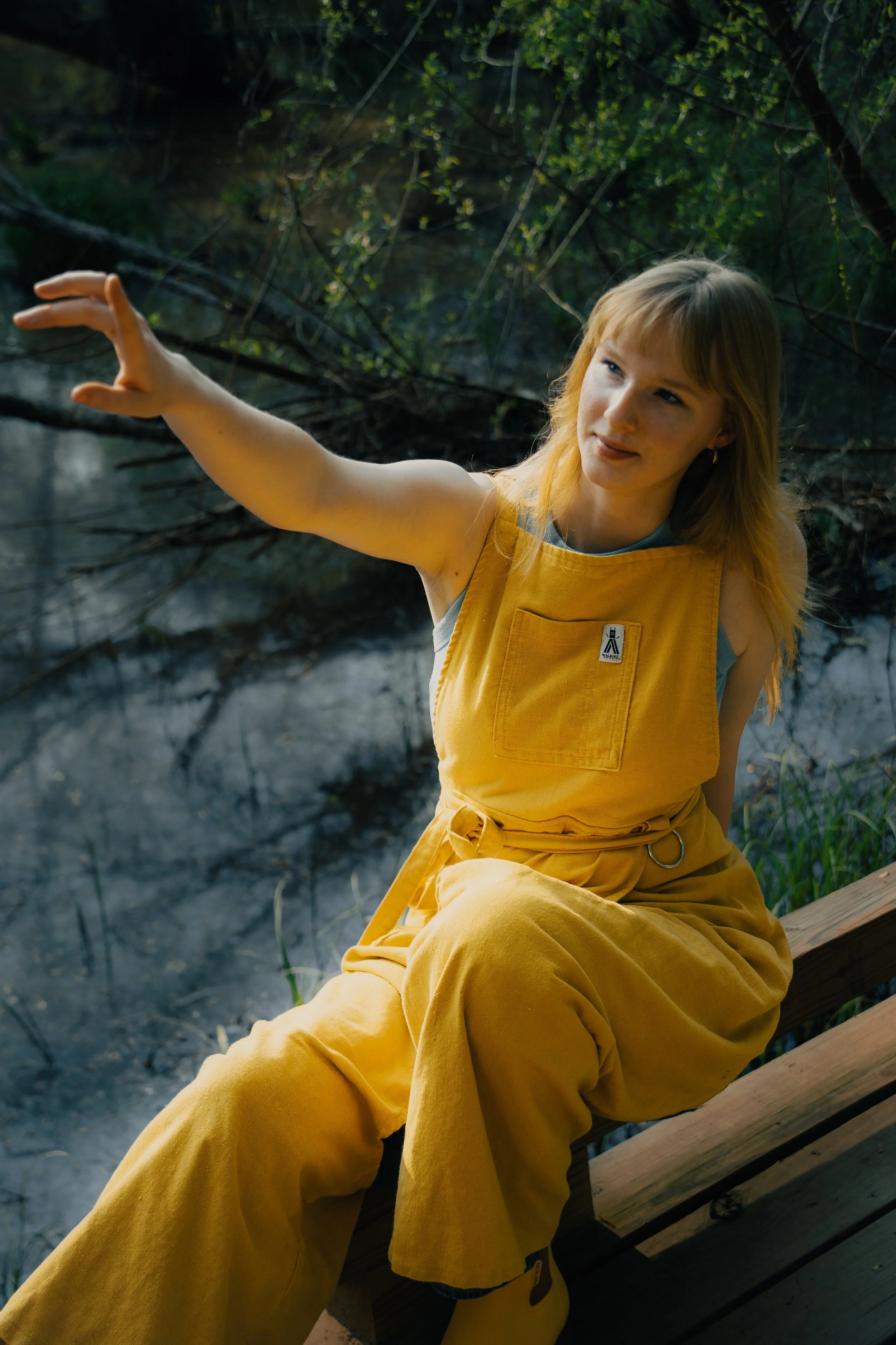 A woman wearing a yellow jumpsuit is sitting on a wooden dock near a river, reaching out her arm towards the water with a focused expression.