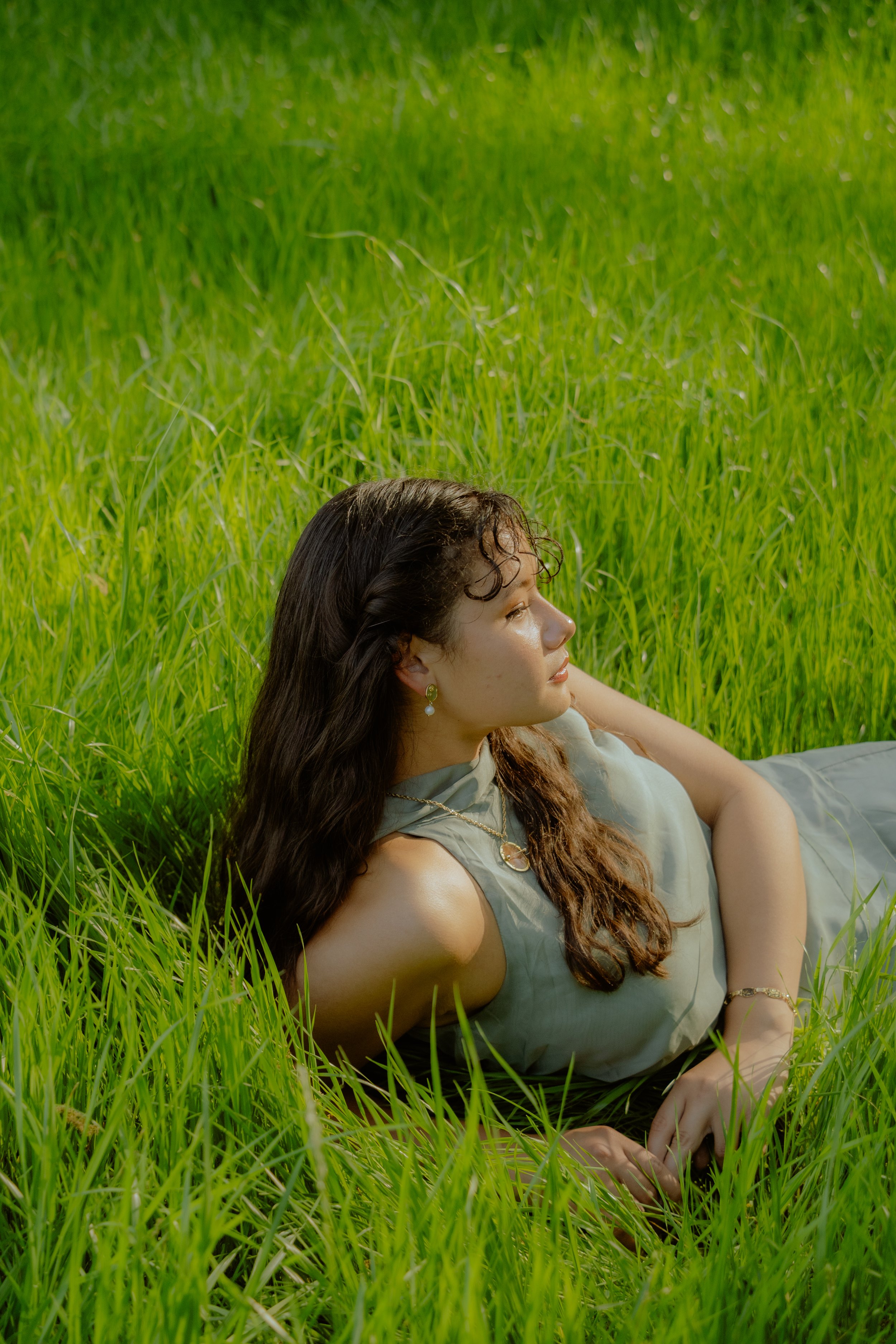 A woman with long dark hair lying in green grass, wearing a sleeveless gray dress and jewelry, looking to the right.