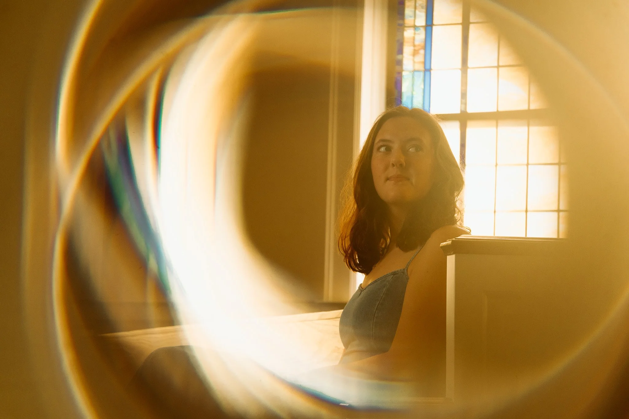 A woman with dark, wavy hair sitting on a bed, looking out the window with sunlight streaming through colorful stained glass, viewed through a prism.