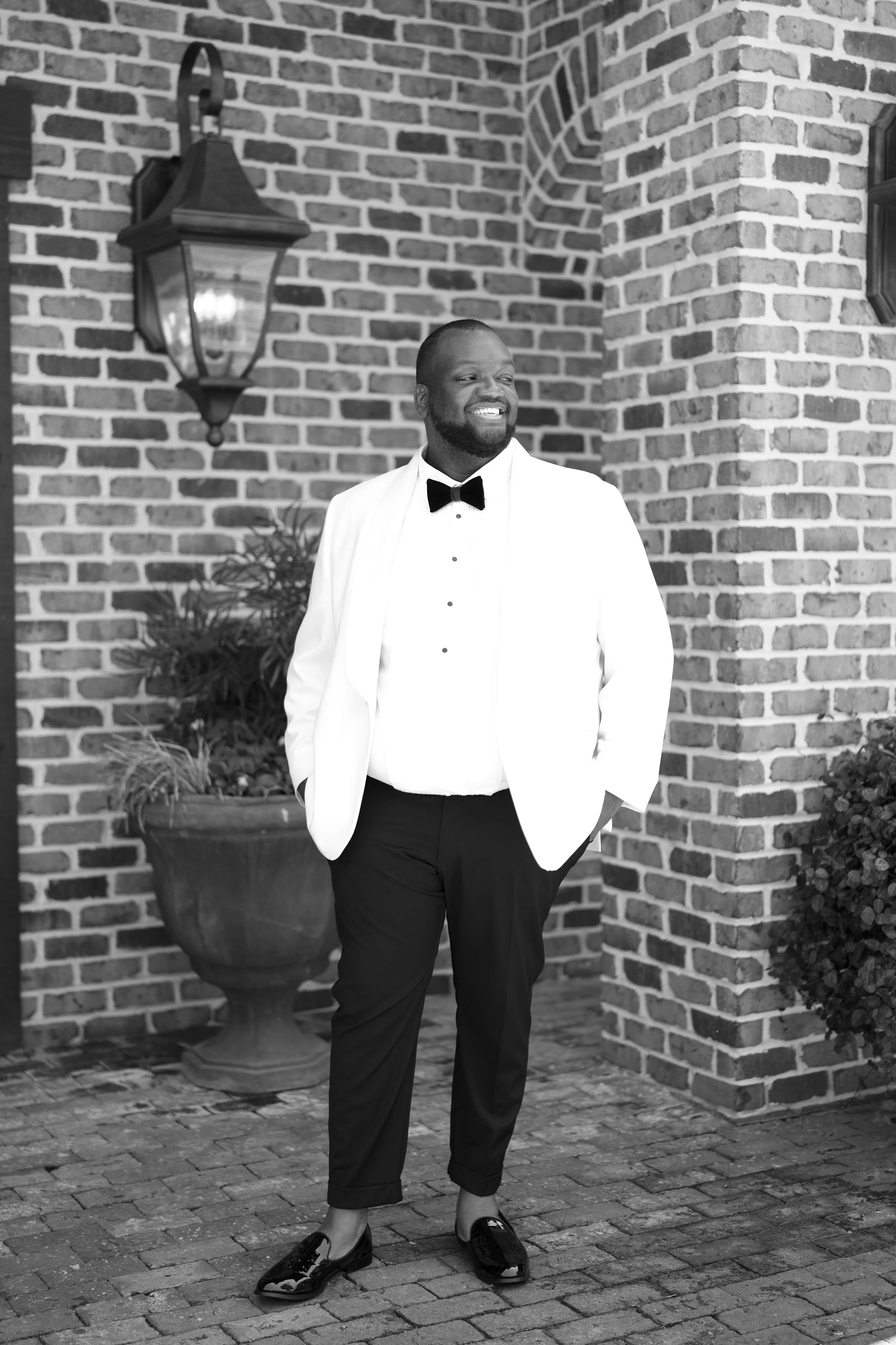 A man dressed in formal attire, including a white tuxedo jacket, black trousers, and a black bow tie, standing outdoors on a brick-paved surface. He is smiling, with his hands in his pockets, and wearing shiny black dress shoes. There is a brick wall with a lamp and decorative plants behind him.