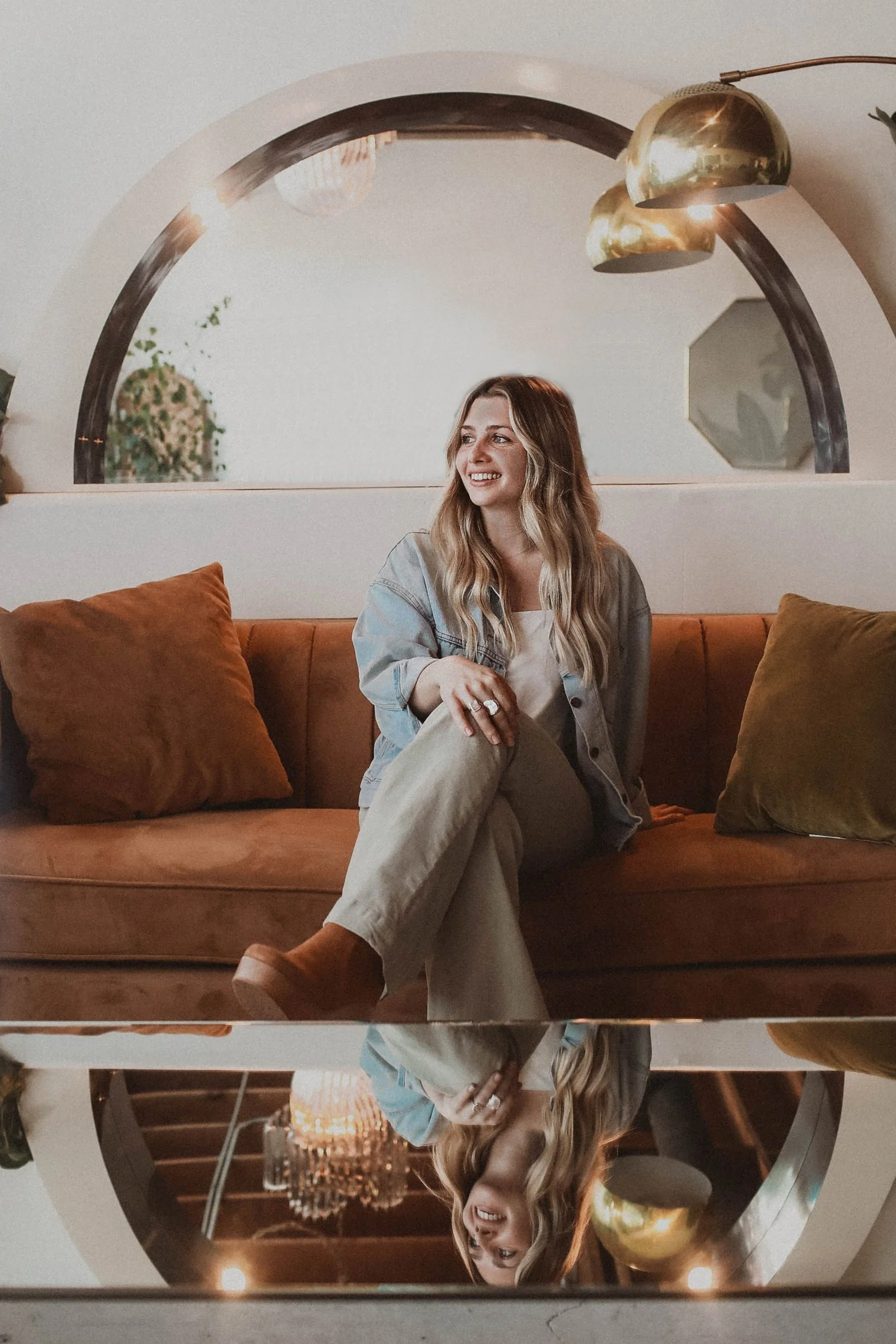 A young woman with wavy blonde hair sitting on a brown sofa with a glass of wine in her hand, smiling, in a modern living room with a large round mirror behind her, reflecting her and the room decor including a chandelier and a decorative vase.
