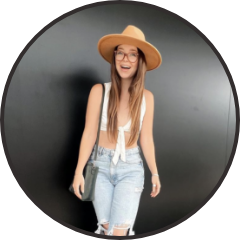 Young woman wearing a large straw hat, glasses, a white sleeveless top, and ripped jeans, standing in front of a dark background and smiling.