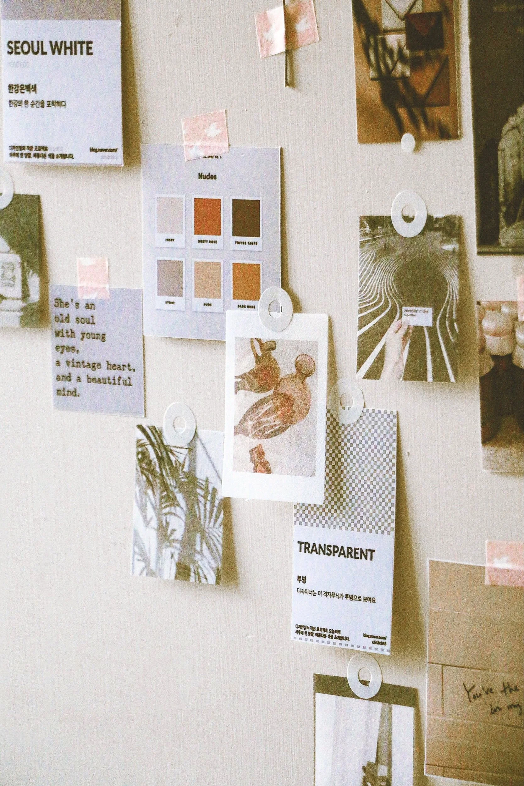 A wall decorated with various printed images and notes, including a color palette labeled 'Nudes,' a quote about someone with young eyes and a vintage heart, and a piece about transparency, among other photos and paper pieces.