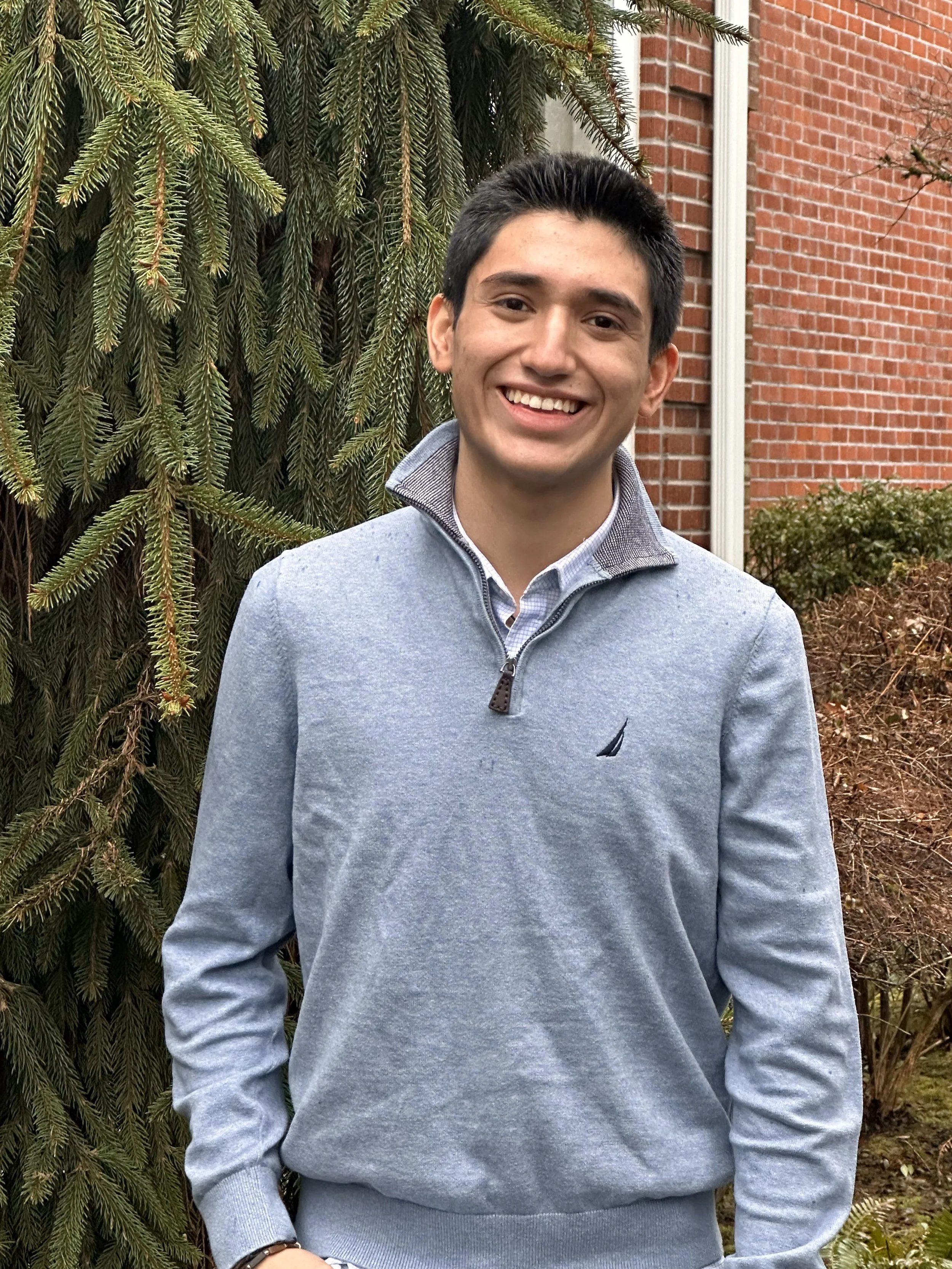 Smiling young man standing outdoors in front of a pine tree and a brick building, wearing a light grey zip-up sweater over a collared shirt.