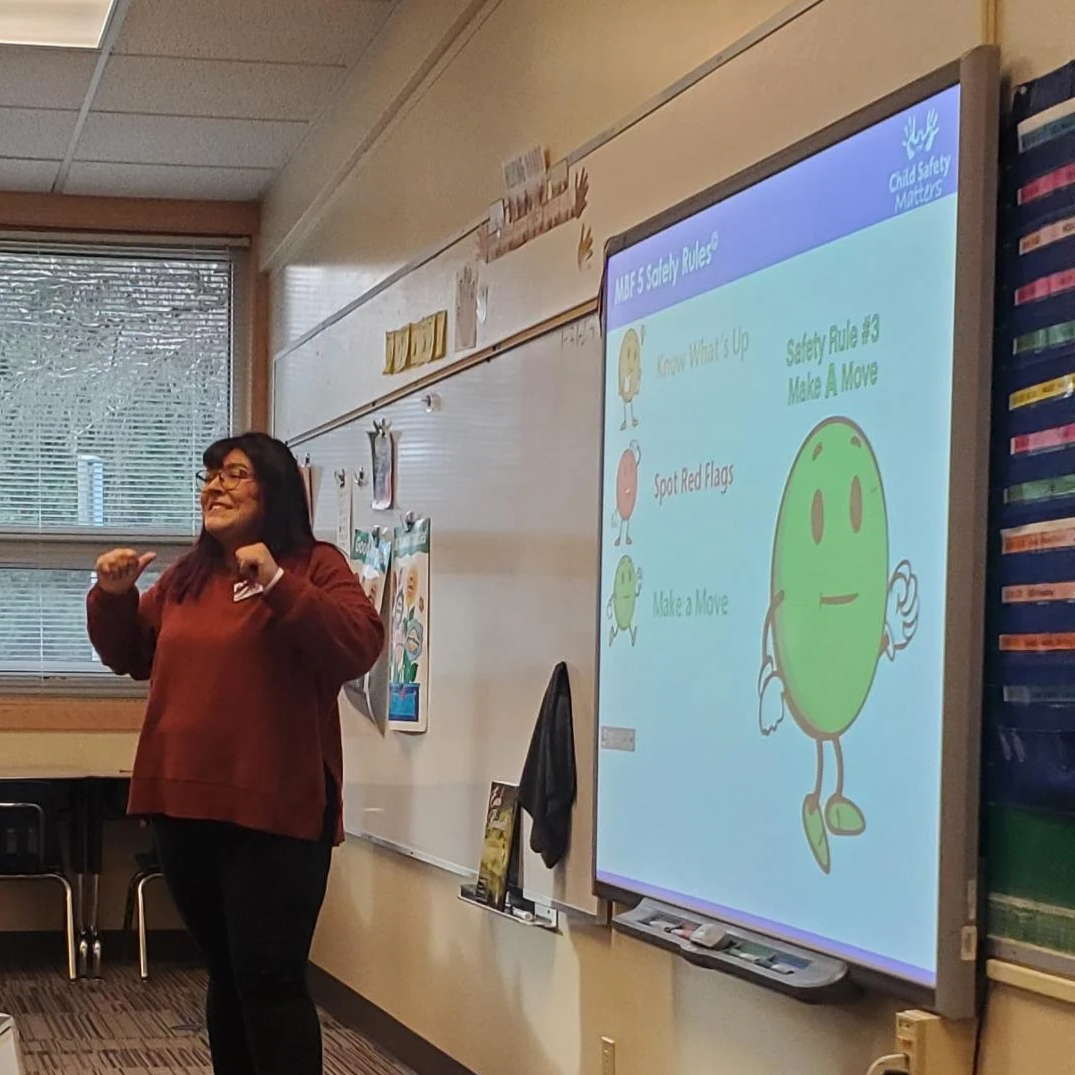 Teacher standing in front of a classroom whiteboard with a projected slide about child safety rules, smiling and wearing glasses and a maroon sweater.