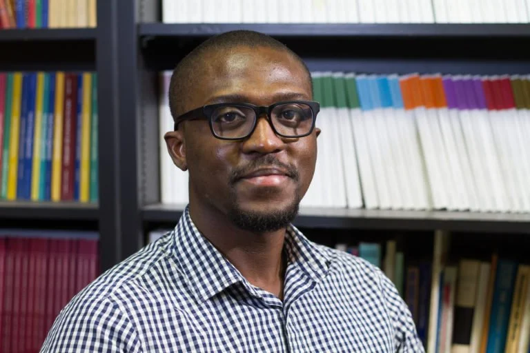 Portrait of a man with glasses and a checked shirt standing in front of a bookshelf