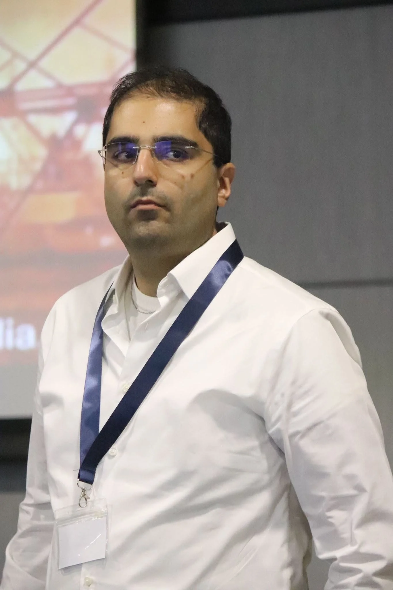 A man wearing glasses and a white shirt with a blue lanyard around his neck, standing indoors with a blurred background.