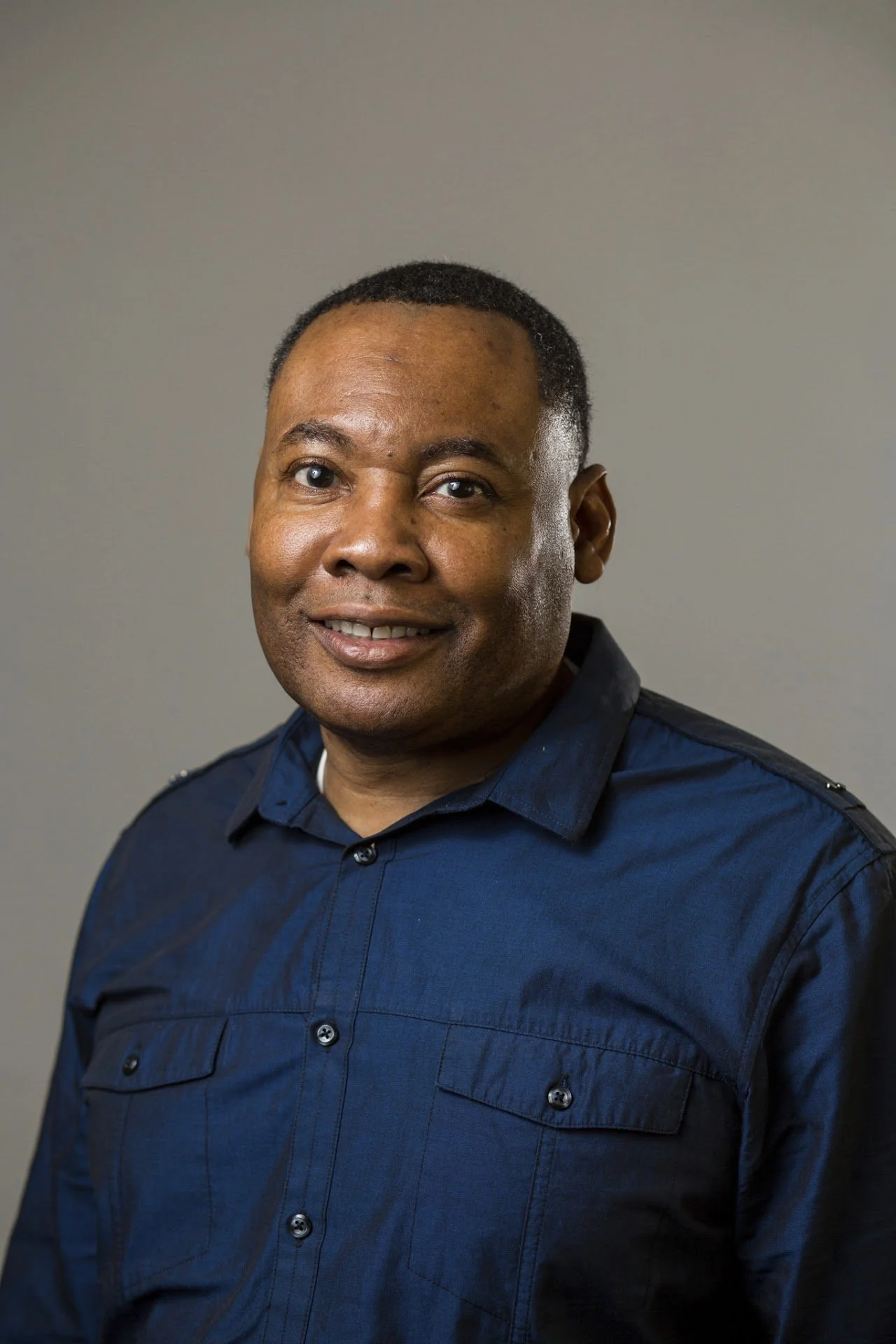 A man with short black hair, dark skin, and brown eyes wearing a navy blue button-up shirt with two chest pockets, standing against a plain gray background.