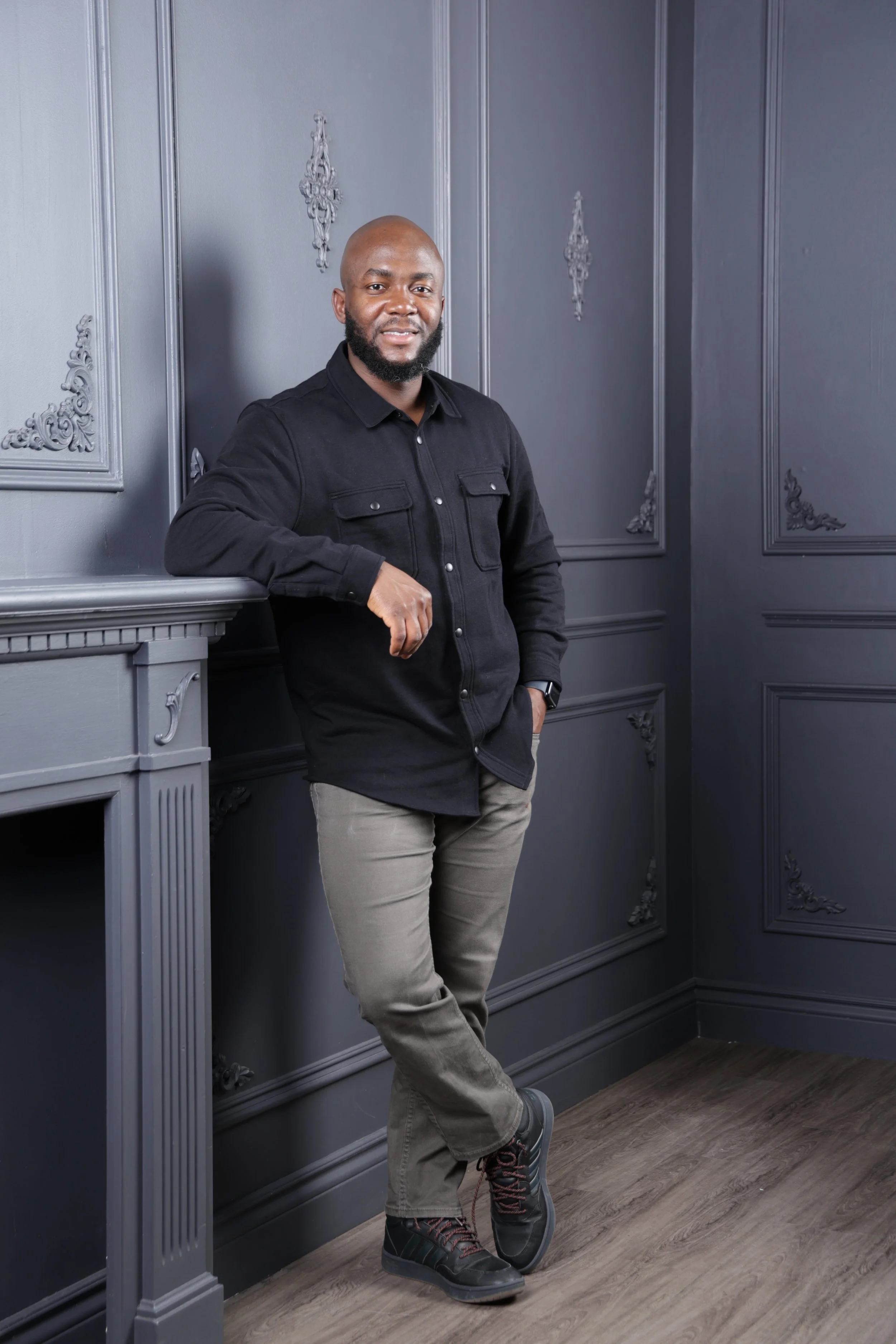 Man in black shirt and gray pants leaning on a mantel in a dark gray room.