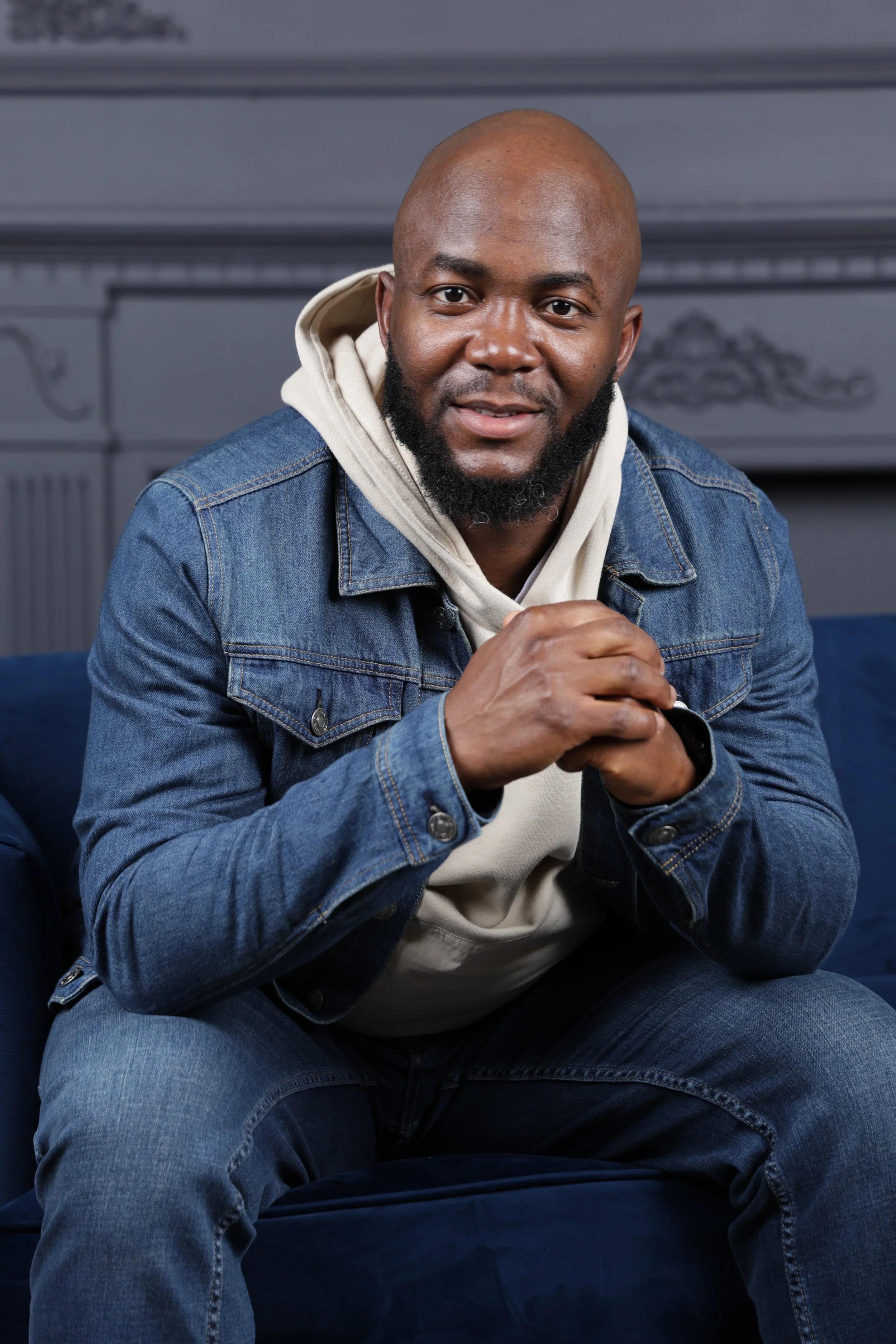 A man wearing a denim jacket and a hoodie, sitting on a blue couch, smiling at the camera.