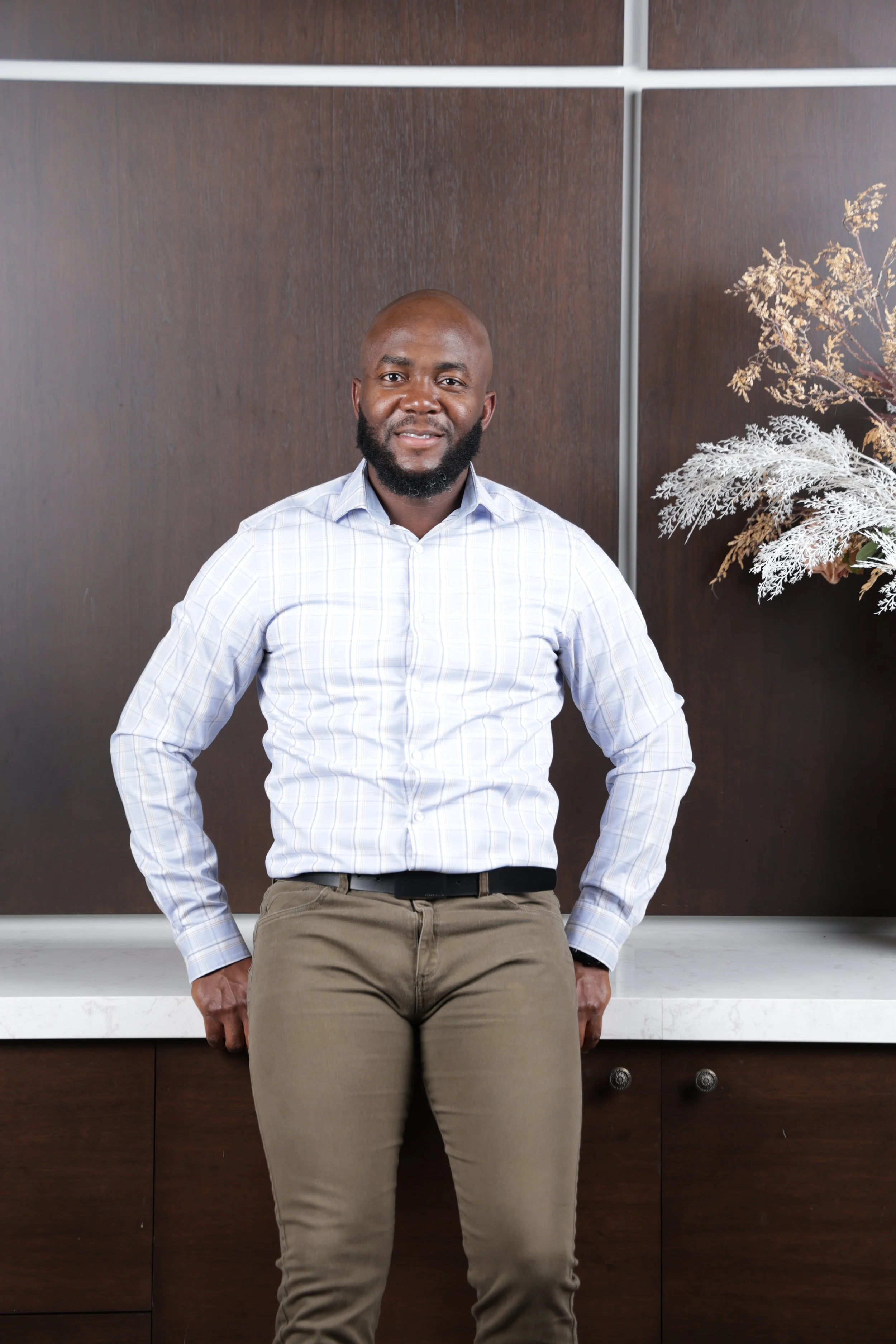 Man in a plaid shirt leaning against a countertop with wooden background and decorative foliage to the side.