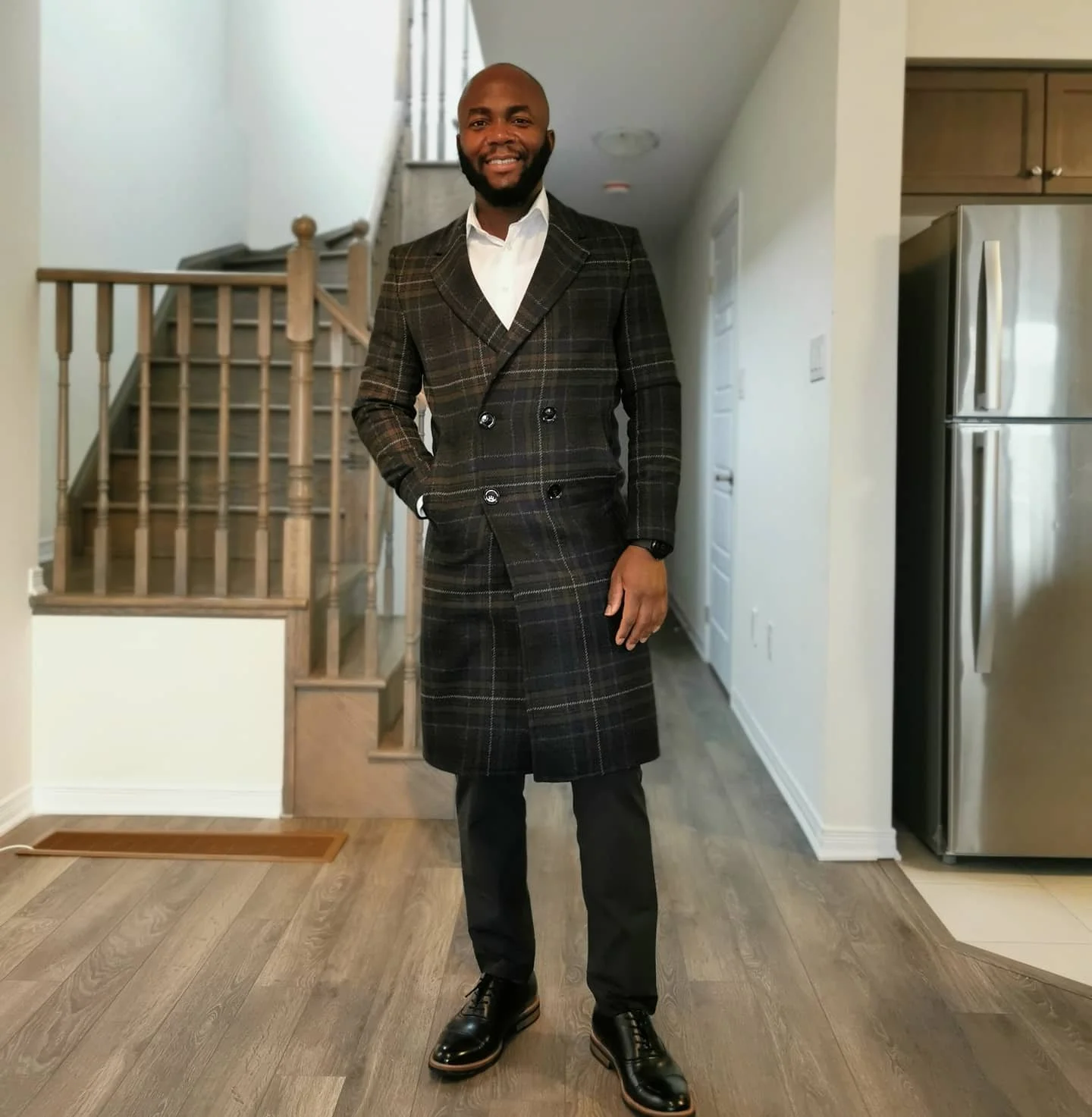 A man in a plaid coat and black pants standing in a modern home interior with wooden stairs and a stainless steel refrigerator in the background.