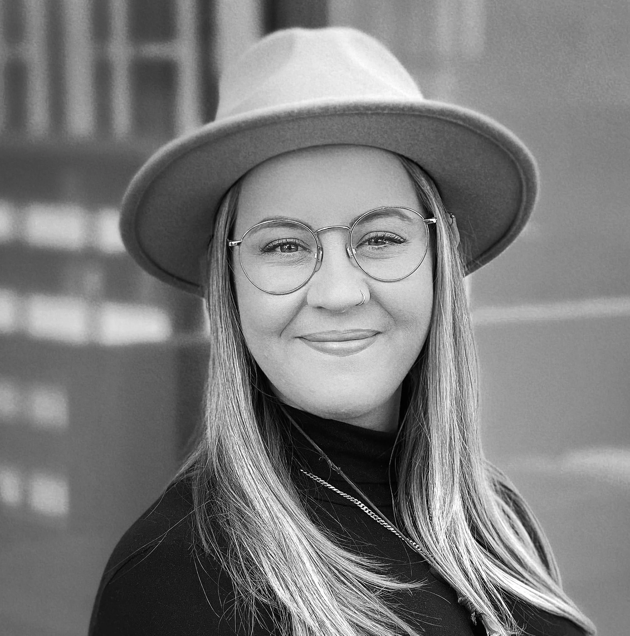 A woman with light brown hair wearing round glasses, a beige wide-brim hat, a black turtleneck, and a silver chain necklace, smiling against a blue background.