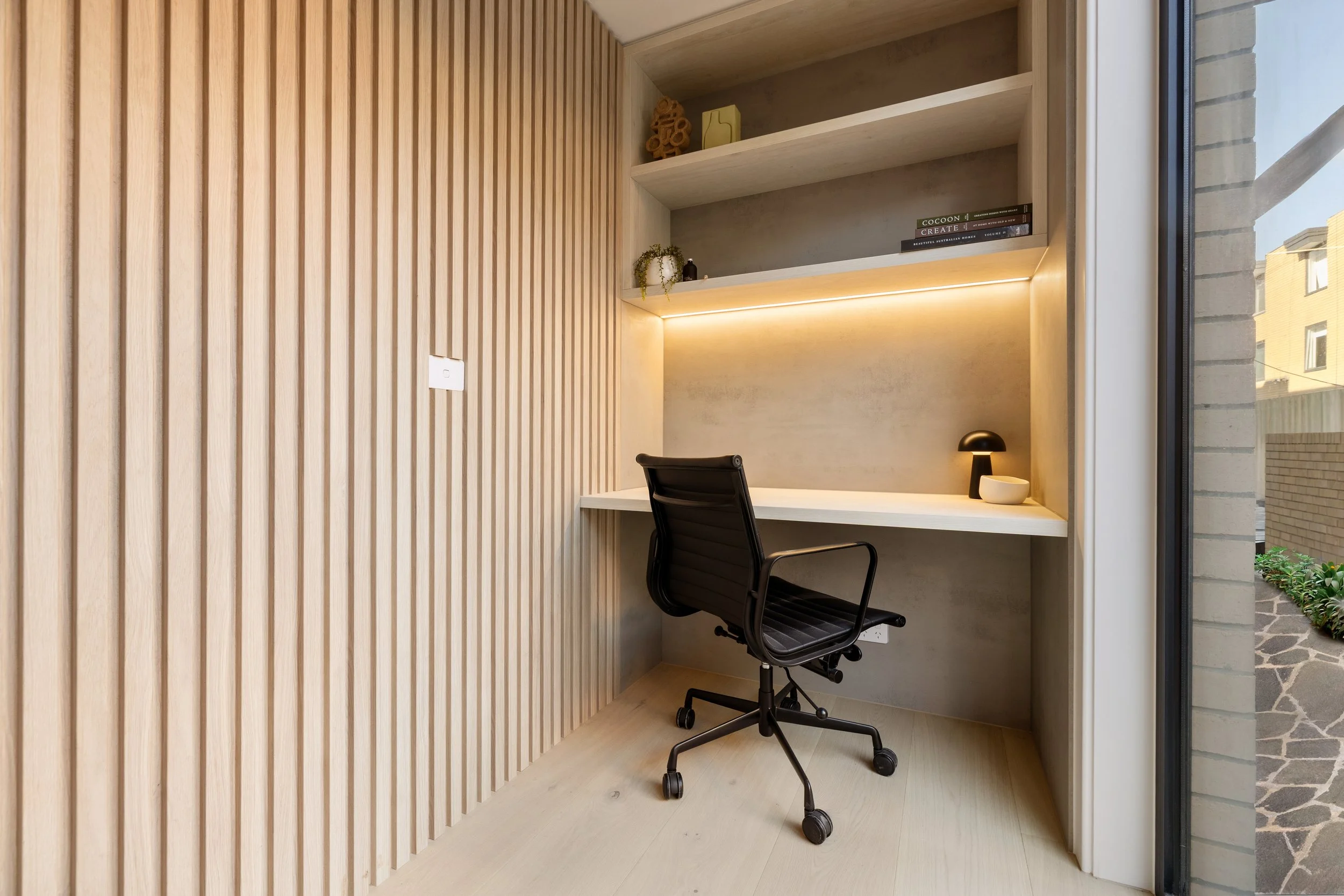 A modern workspace with a white desk, black office chair, built-in wooden shelves, and a window showing neighboring buildings and a garden.