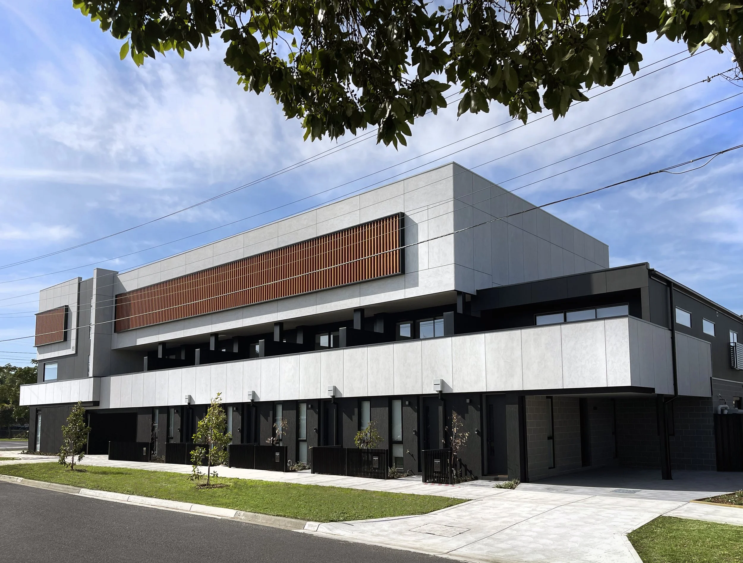 Modern building with white and black exterior walls, large windows, and wooden slats on upper levels, situated next to a sidewalk with small trees, under a blue sky with scattered clouds.