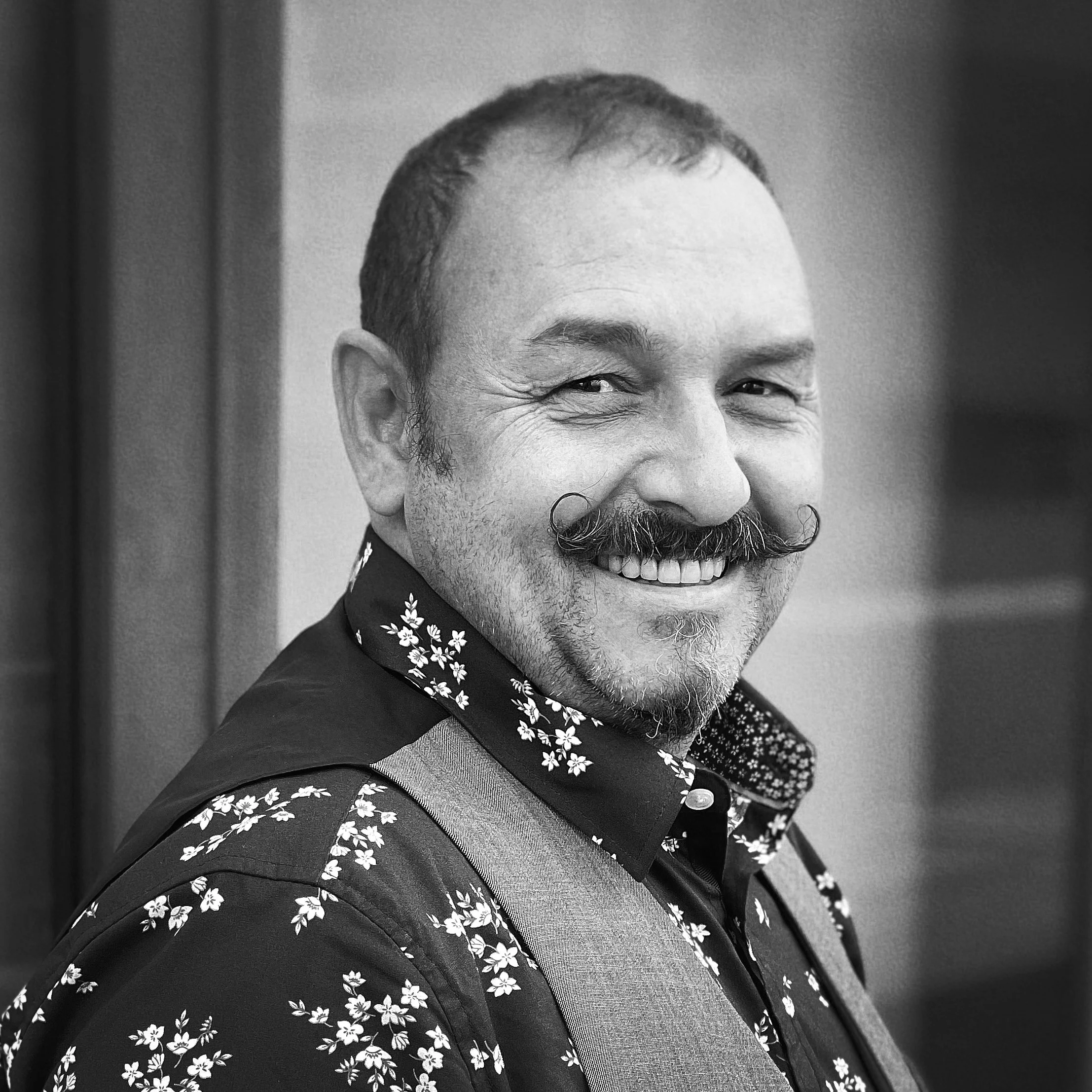 Close-up of a smiling Asian man with short hair, mustache, and beard, dressed in a dark jacket with a floral pattern shirt underneath.