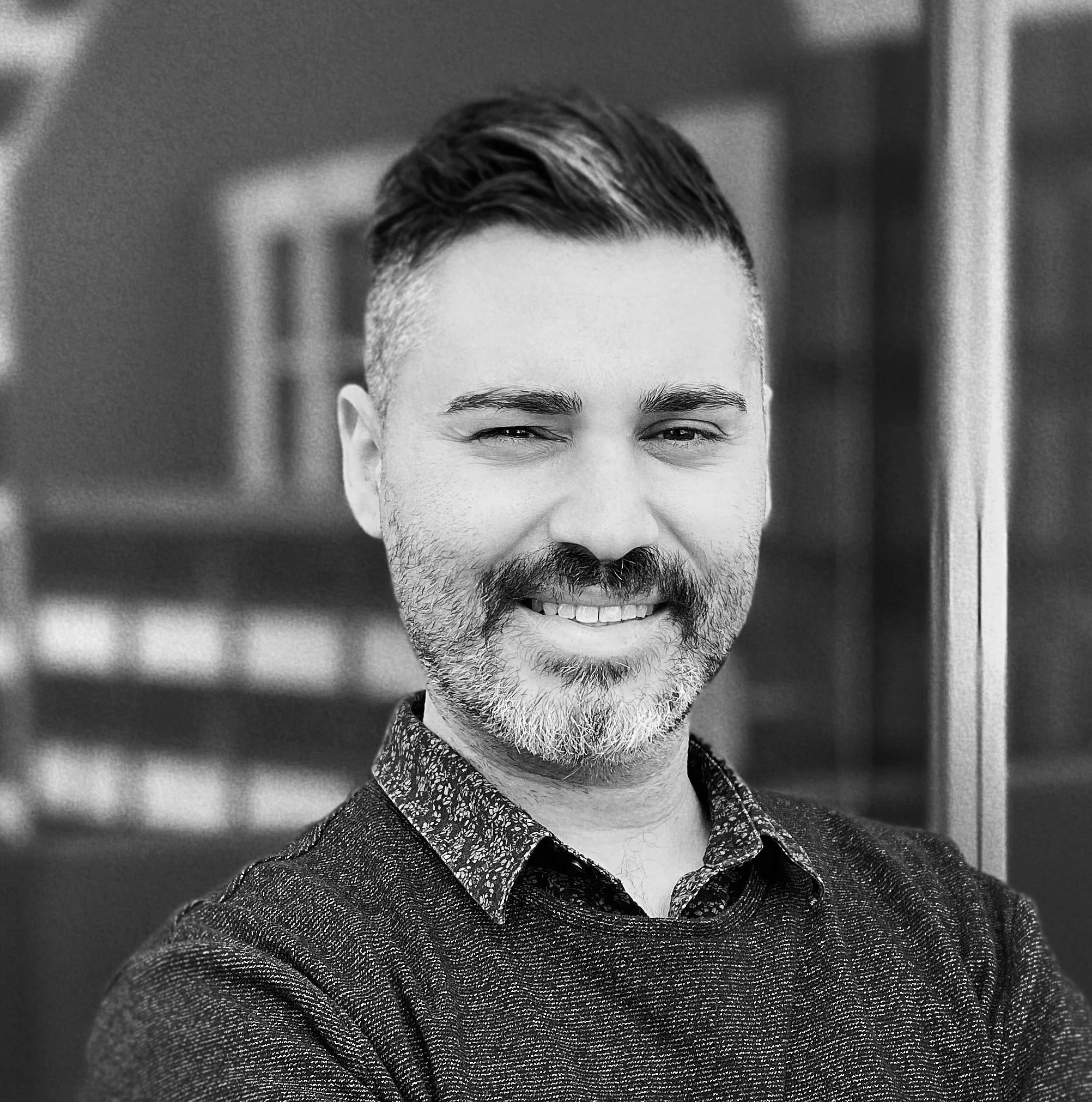 Portrait of a smiling man with dark hair, beard, and mustache, wearing a dark sweater over a patterned shirt, standing outdoors in front of a blue building.