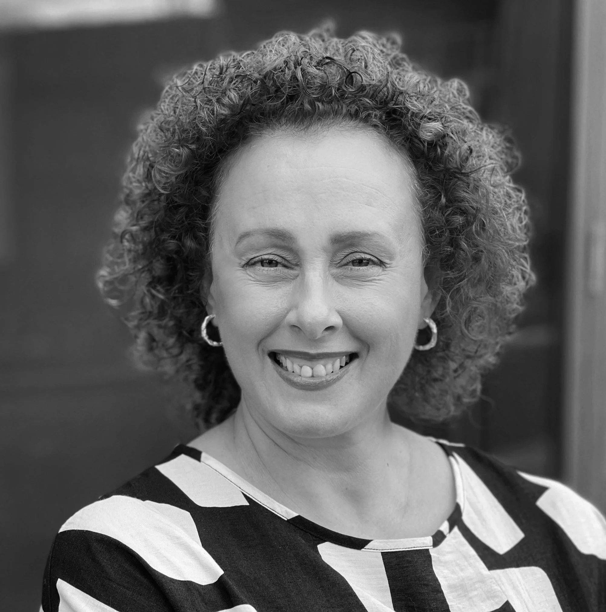A woman with curly red hair smiling, wearing hoop earrings and a black and white geometric patterned top, standing in front of a blue background.