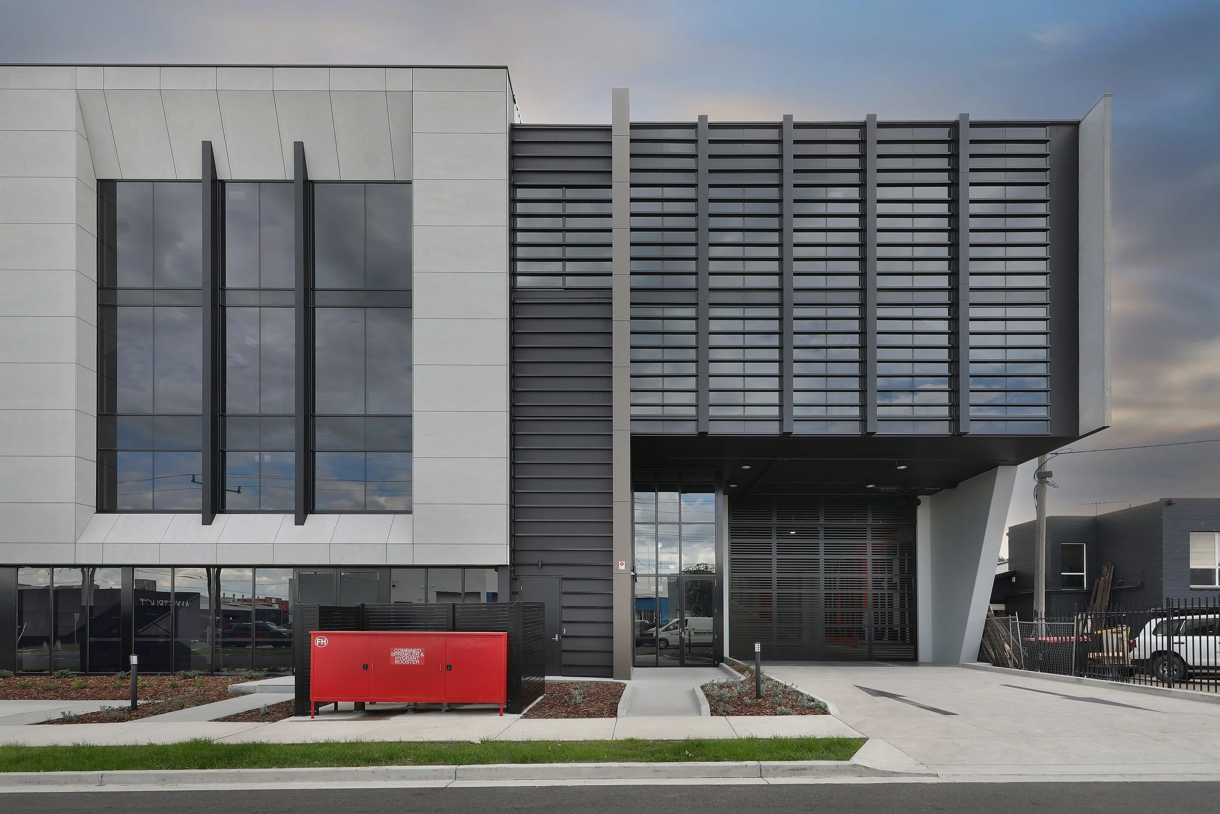 Modern multi-story building with large glass windows, metal louvers on one side, and a concrete sidewalk in front. There is a red fire safety cabinet and some landscaping with grass and small plants near the sidewalk.